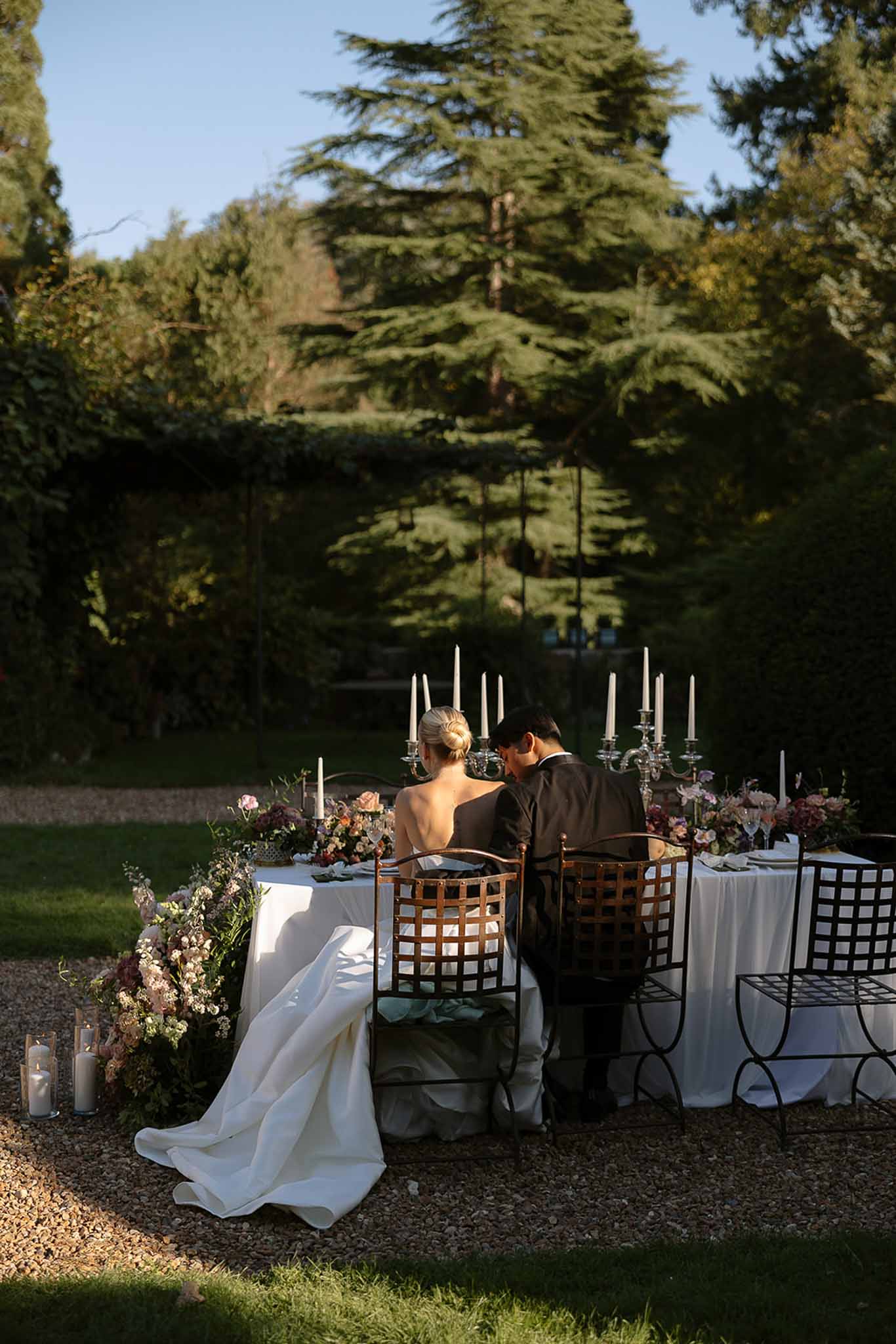 Couple from behind at sweetheart table with silver candelabras and mauve roses in formal garden