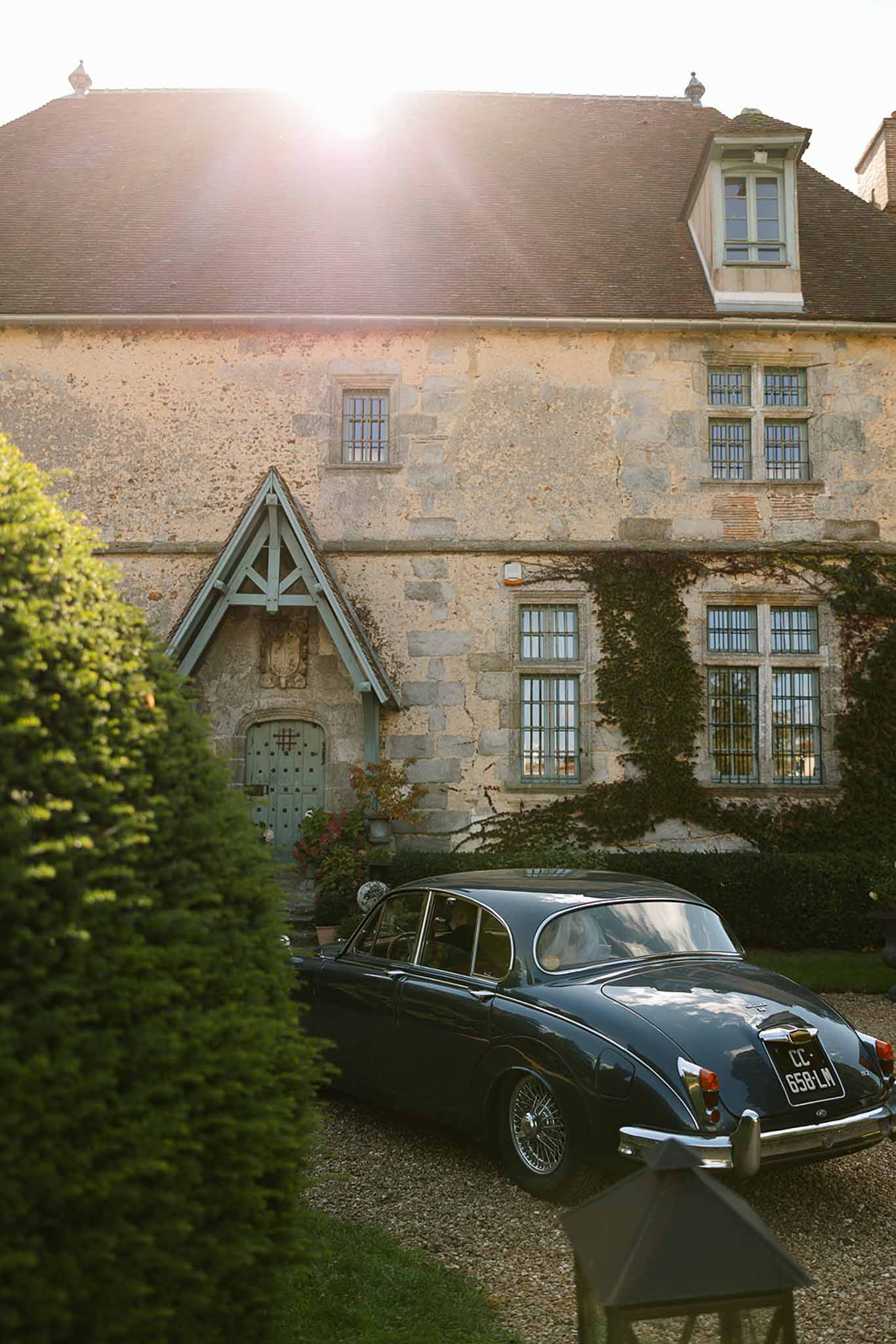 French chateau with limestone walls and slate roof at golden hour with navy vintage Jaguar parked on gravel driveway
