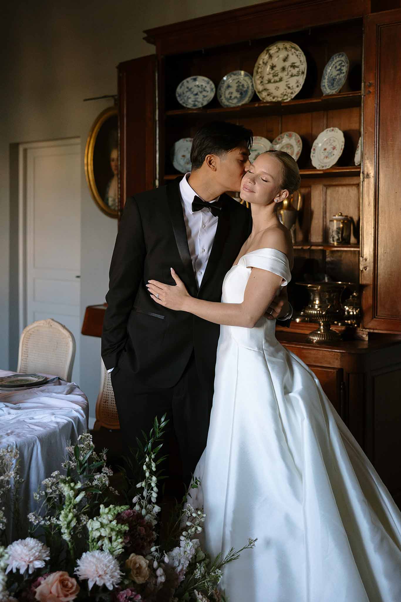 Groom kissing bride temple beside blush rose arrangement with antique cabinet and portrait behind