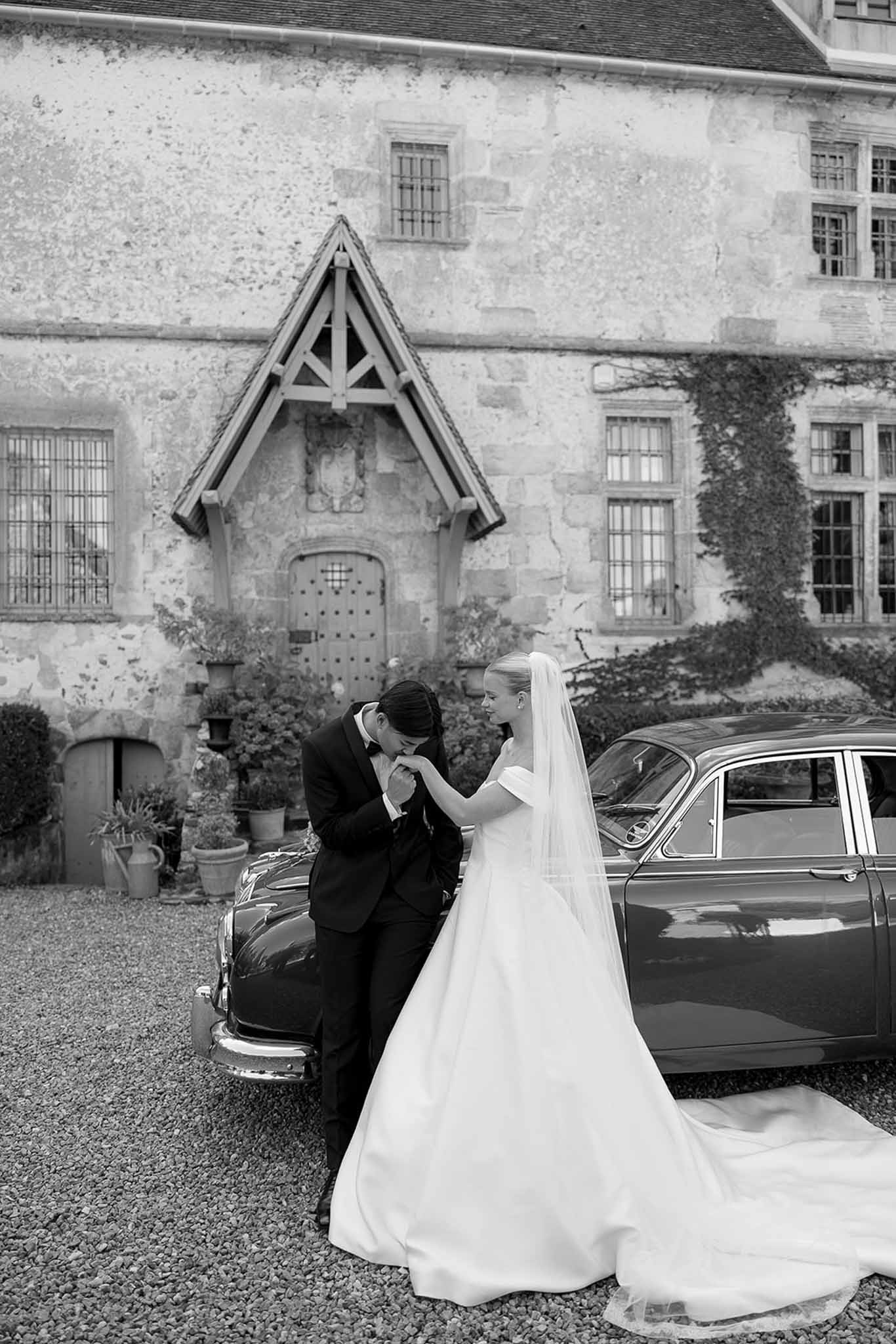 Black and white groom kissing bride's hand beside vintage car before ivy-covered chateau with pointed porch