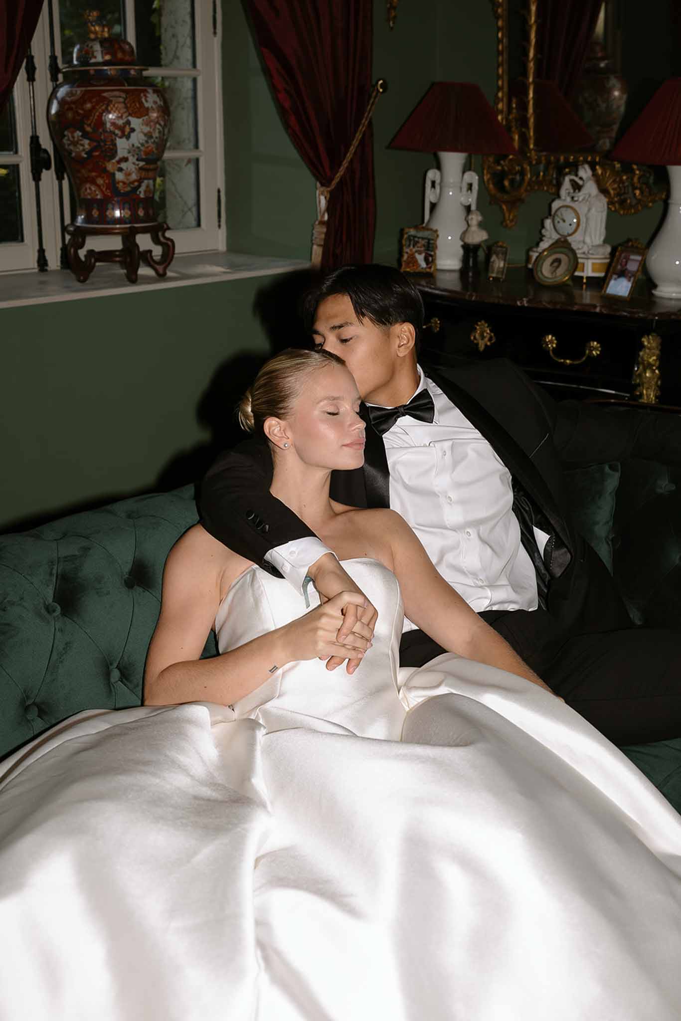 Groom kissing bride's temple as they sit on green velvet sofa in richly decorated chateau interior