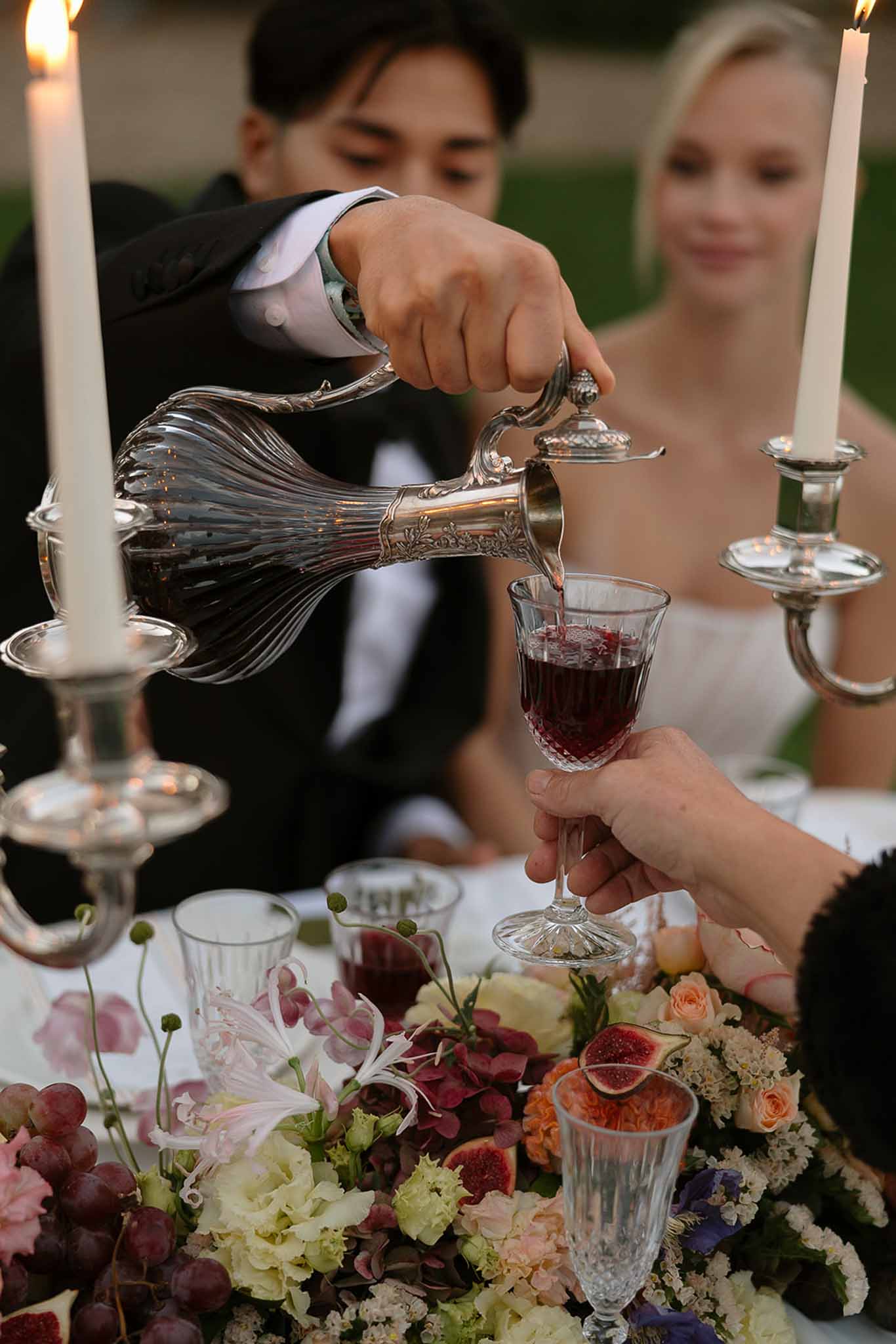 Groom pouring red wine from silver decanter beside jewel-toned centrepiece with figs and grapes