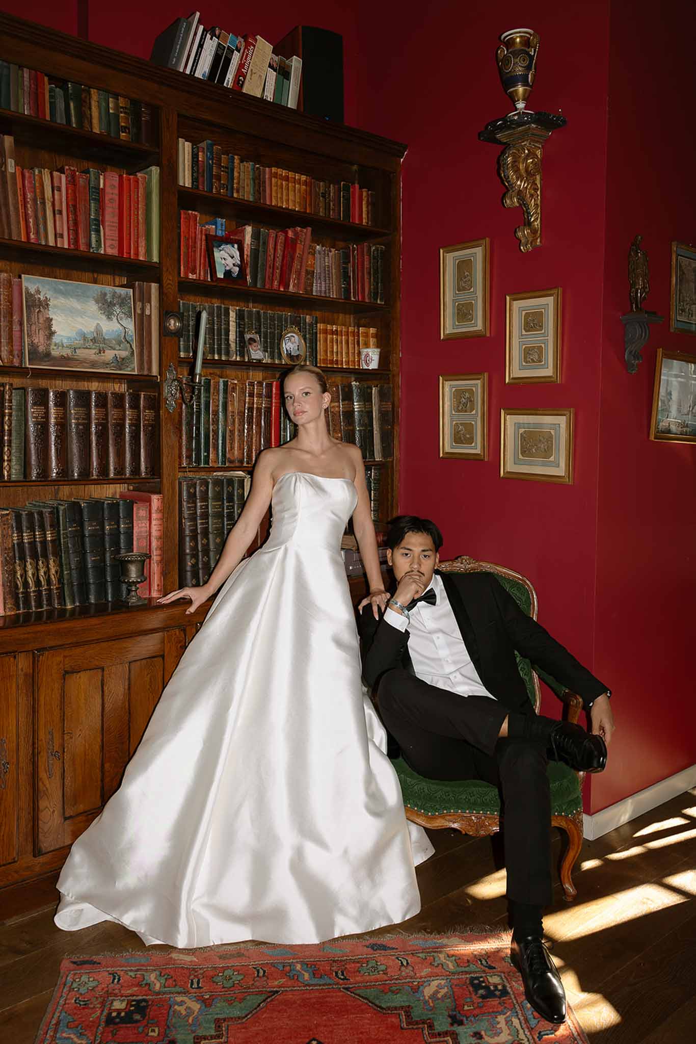 Bride in white ballgown standing beside groom seated in velvet chair in crimson library with bookshelves