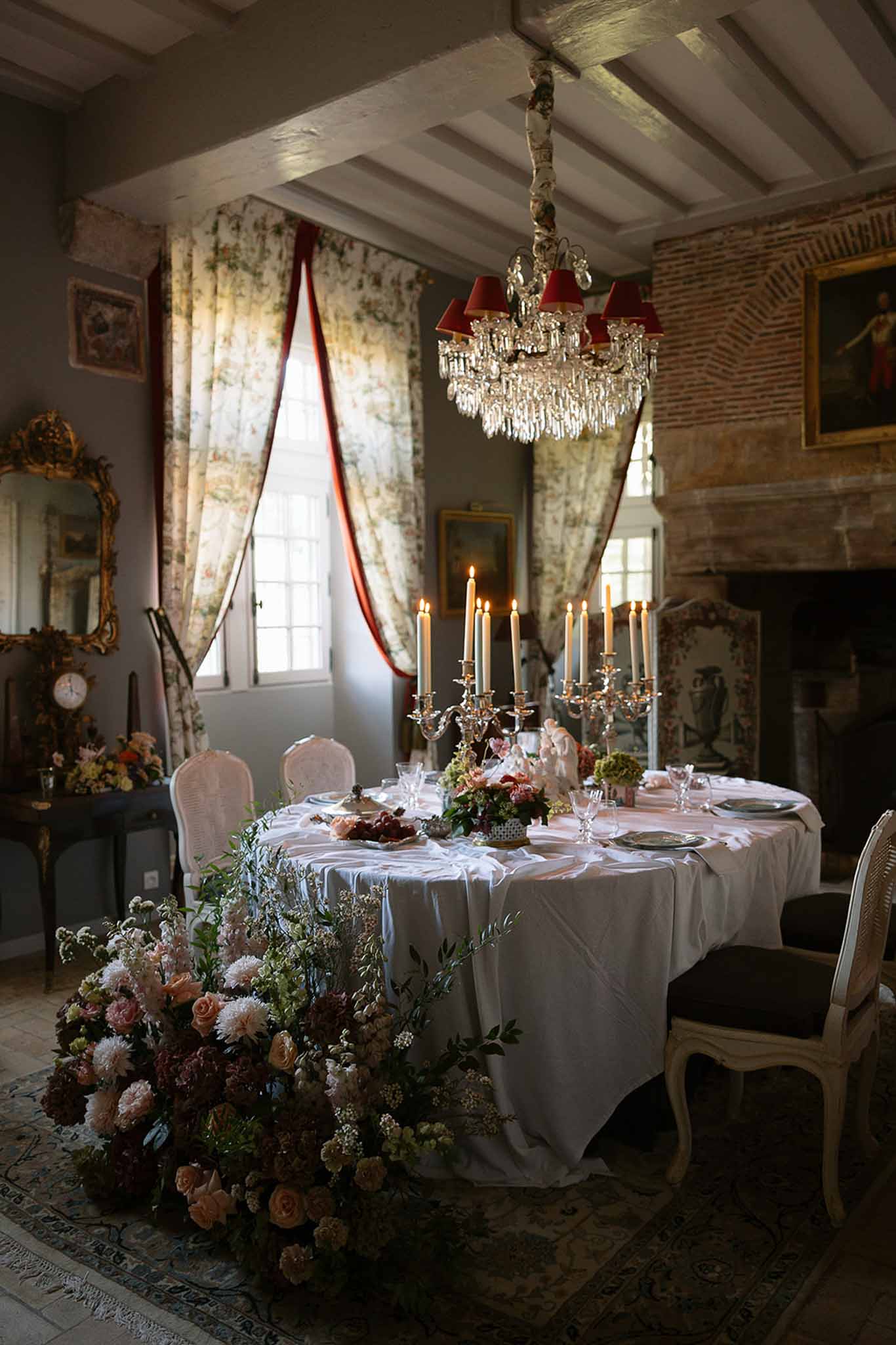 Oval table with blush roses, burgundy ranunculus, and silver candelabra in beamed chateau dining room