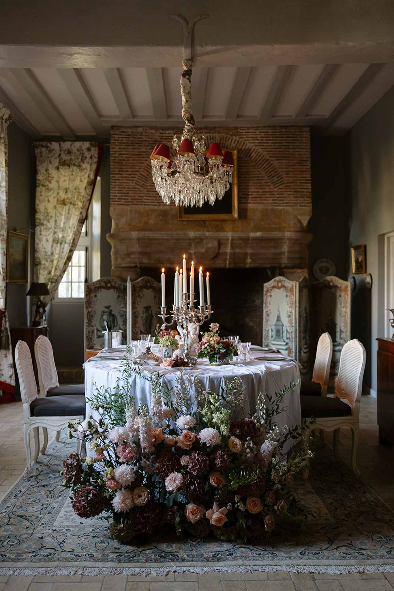 Oval table with cascading blush and burgundy floral arrangement beside brick fireplace in moody candlelit chateau room