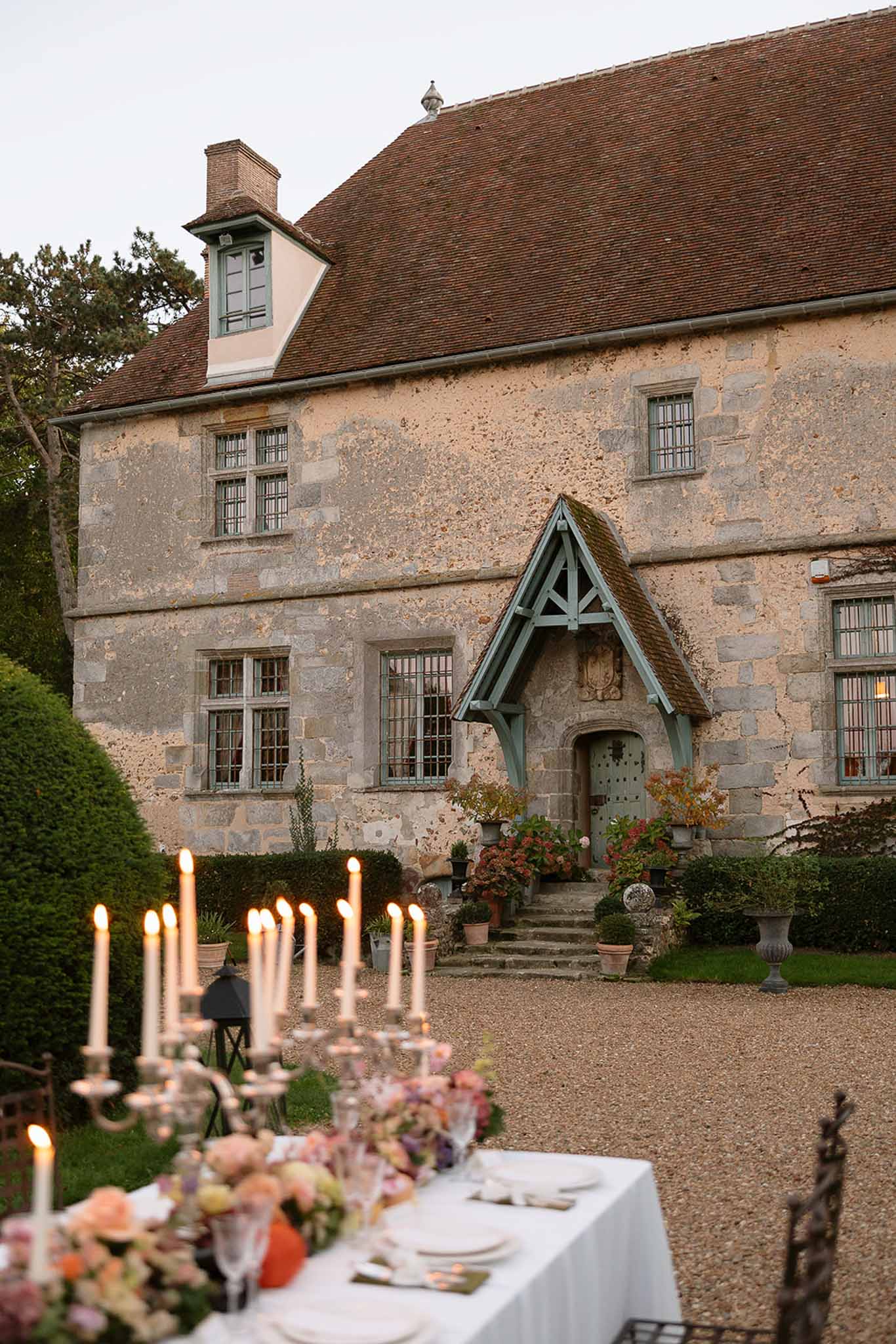 Silver candelabra table with peach and coral florals at dusk before Gothic-porched stone manor on gravel courtyard