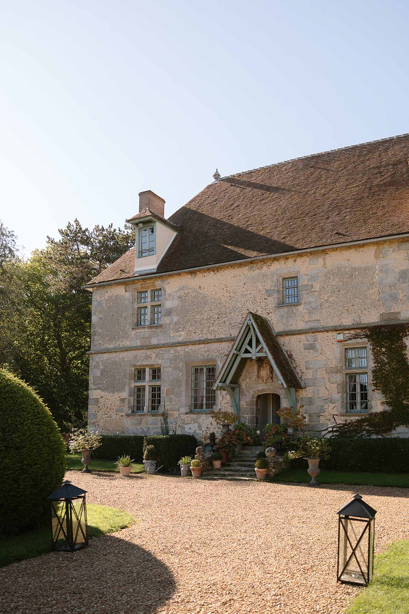 French country manor with limestone facade, blue-painted porch canopy, gravel drive flanked by lanterns and hedging