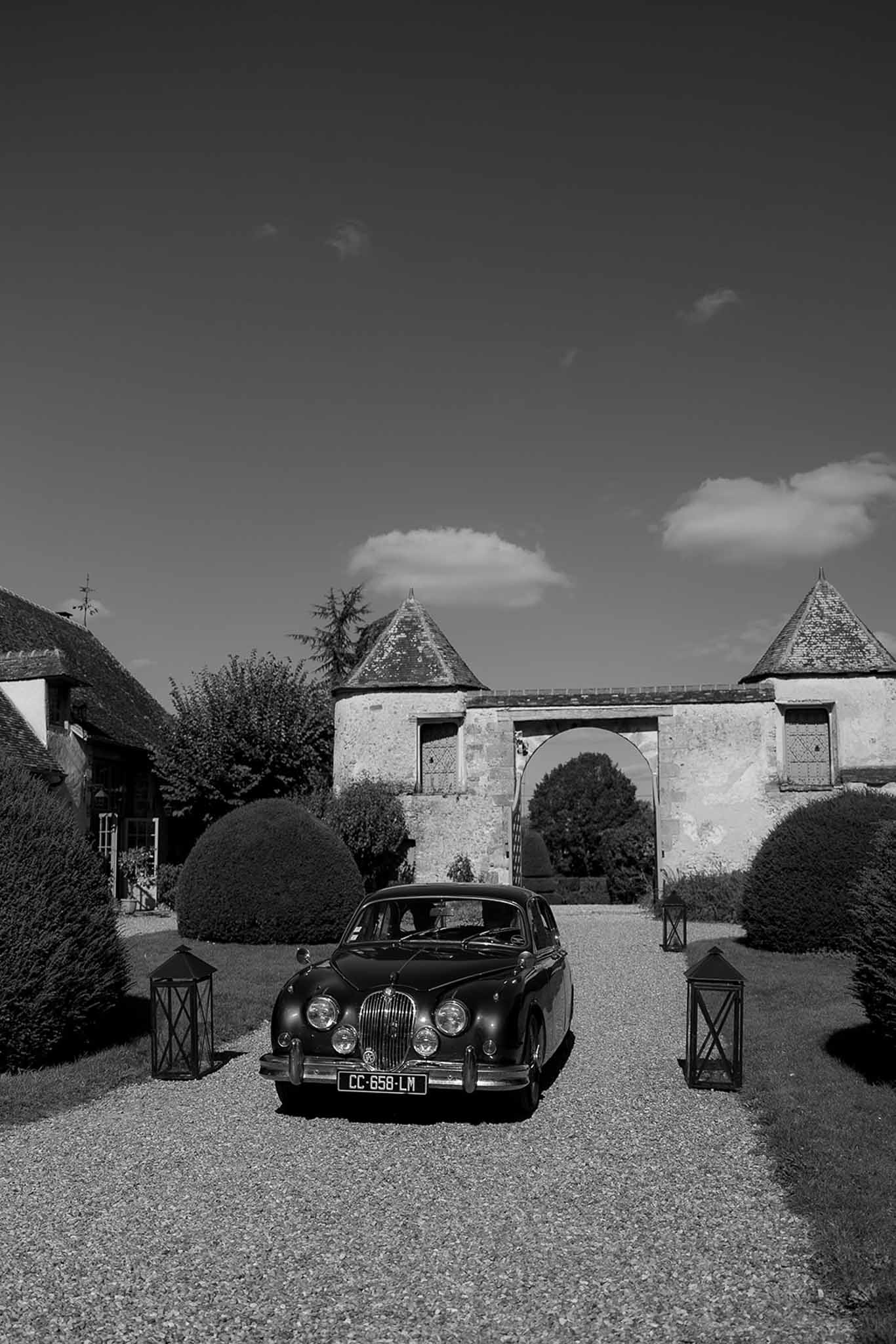 Black and white photo of vintage Jaguar parked before twin-towered chateau gatehouse on gravel driveway