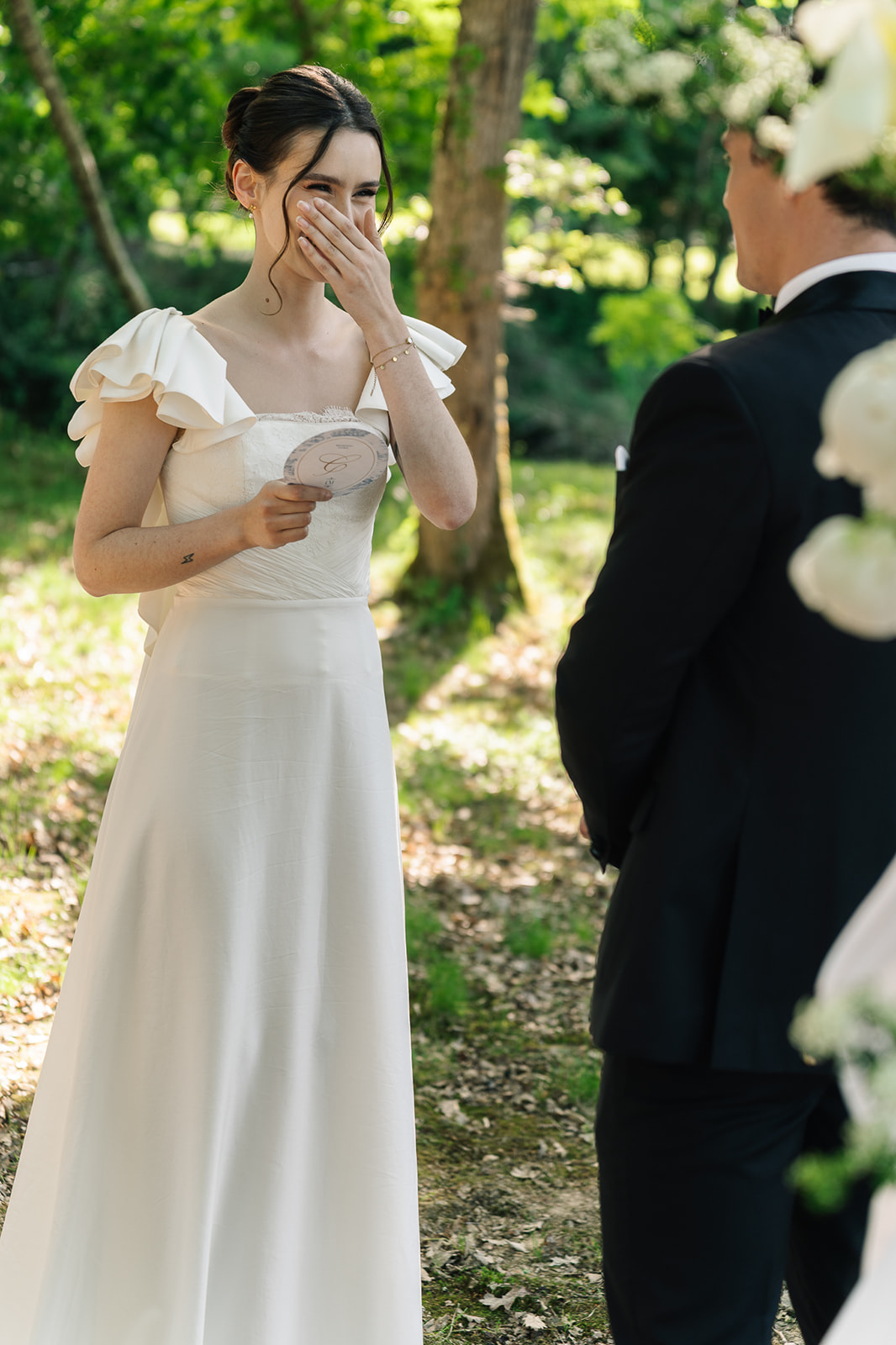 Bride in off-shoulder ruffled gown covers mouth emotionally while reading vows in dappled garden light