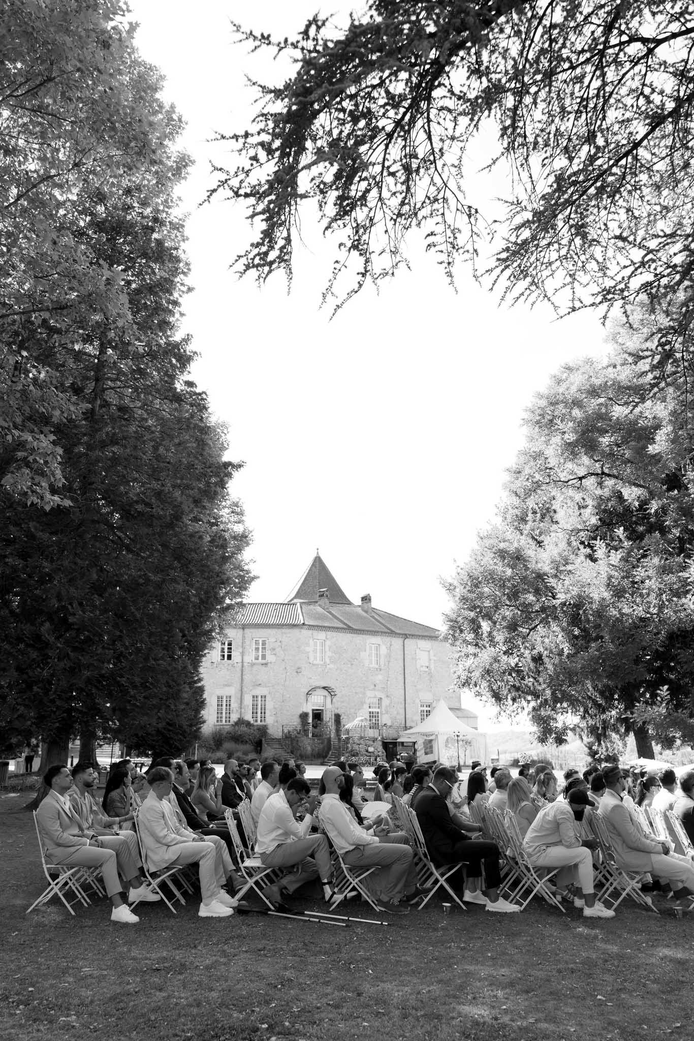 Black and white wide shot of outdoor wedding ceremony with guests seated on lawn facing a French chateau