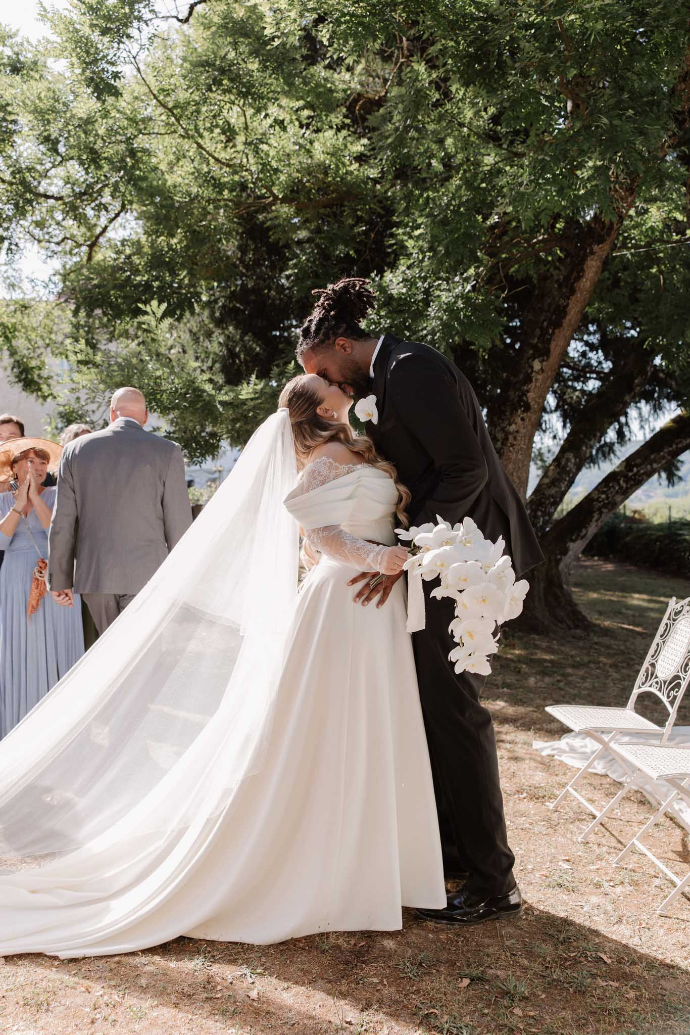 First kiss under tree bride in off-shoulder lace gown with white orchid cascade bouquet groom in black suit