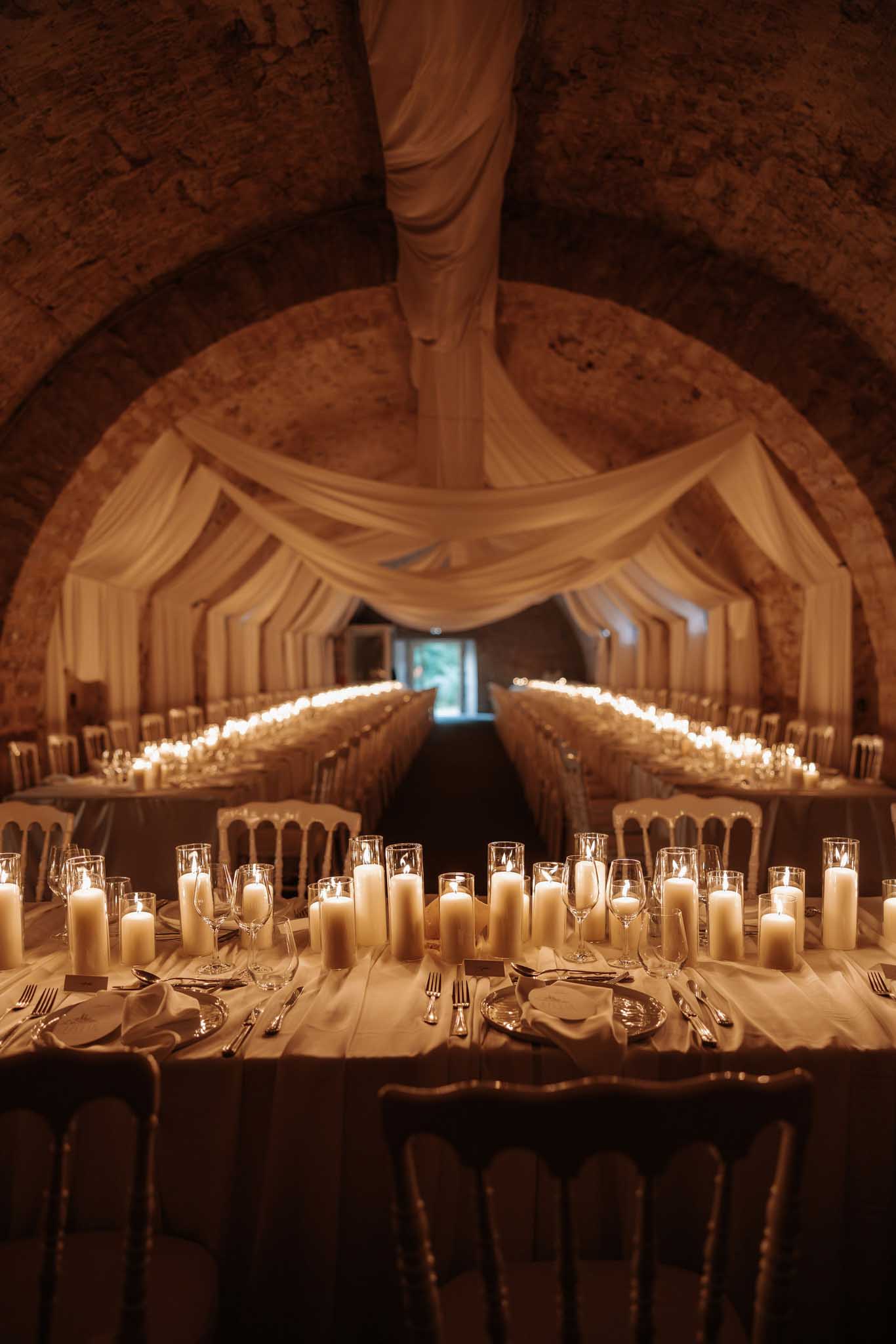 Candlelit vaulted stone cellar with white draped ceiling, ivory runners, pillar candles, and gold Napoleon chairs