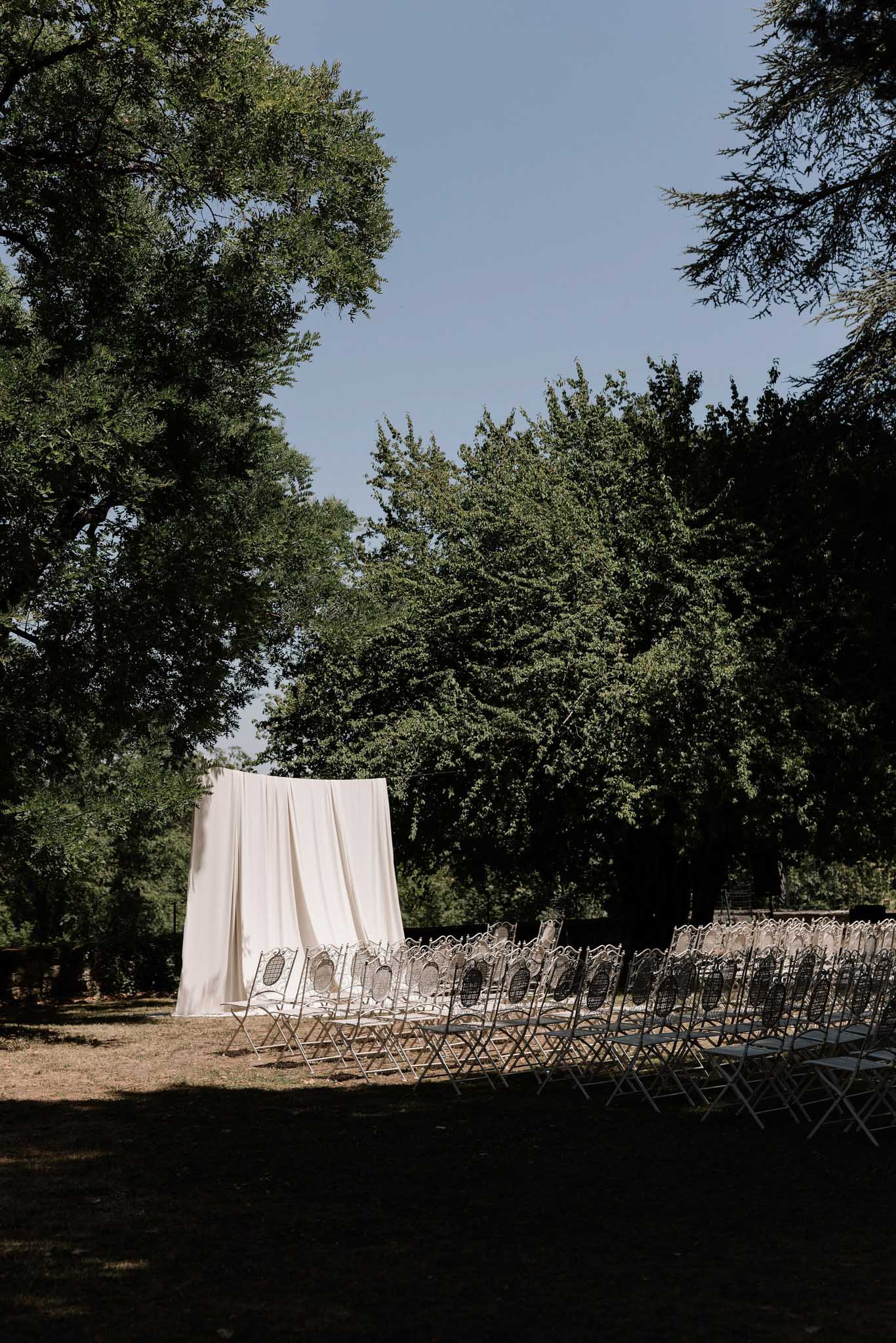 Outdoor ceremony setup with ivory fabric altar backdrop and white ornate metal folding chairs