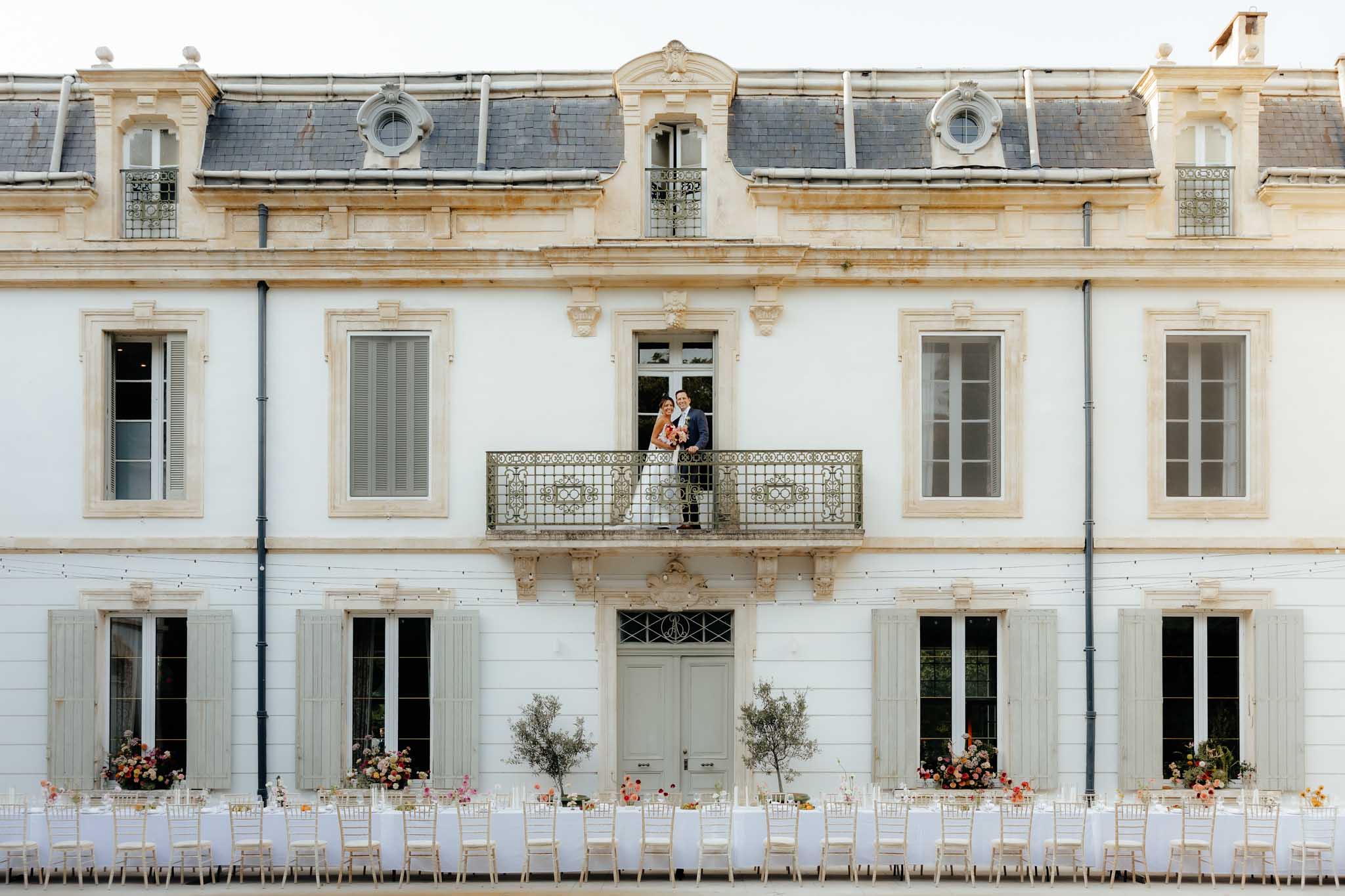 Bride and groom on Juliet balcony of French chateau with outdoor reception tables and fairy lights below