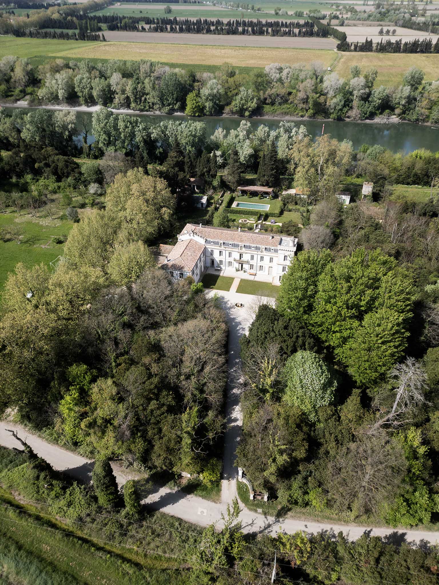 Aerial view of French chateau with terracotta roof, swimming pool, formal gardens, and river alongside