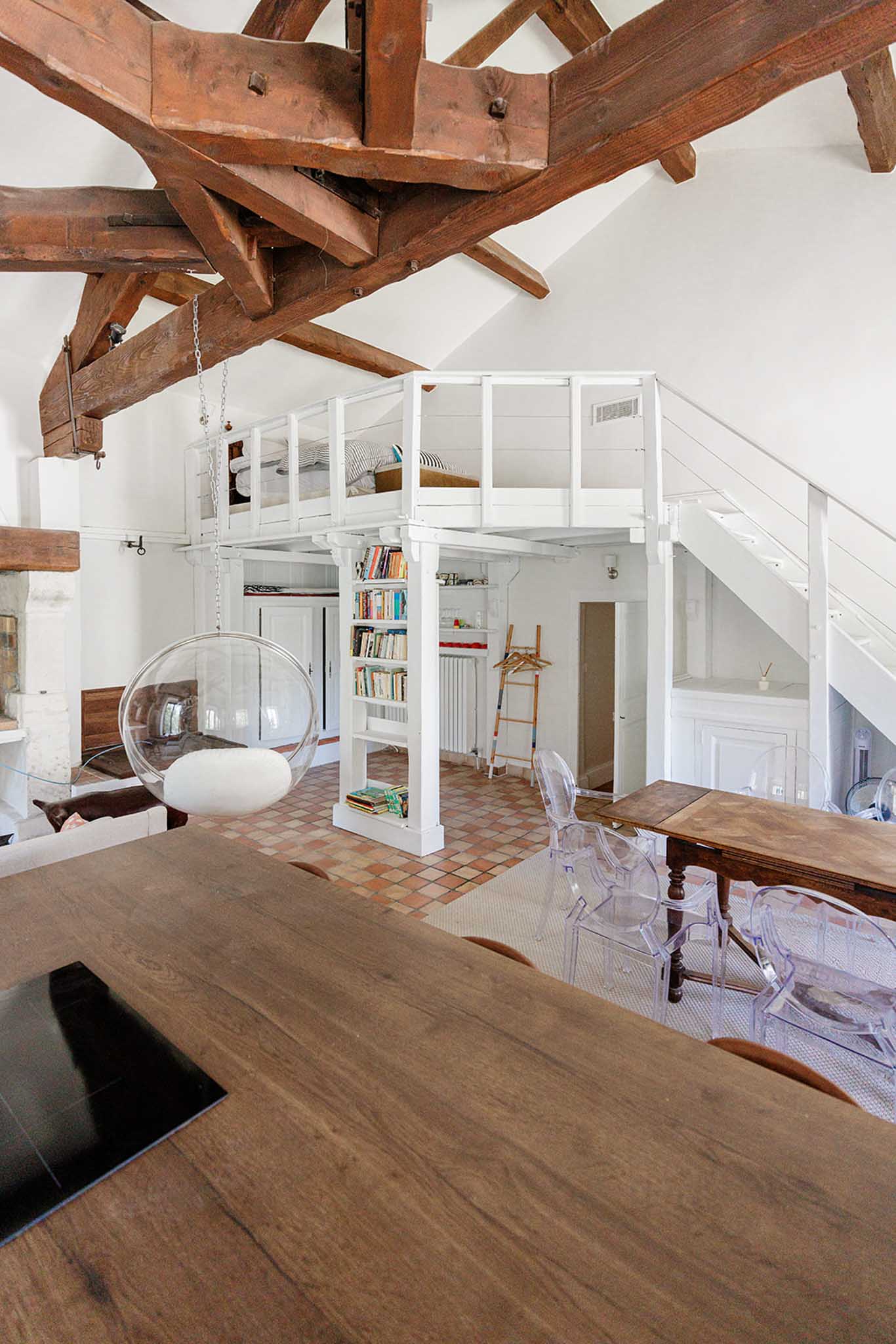 Interior of converted French mill with exposed beams, mezzanine sleeping level, kitchen island, and ghost chairs