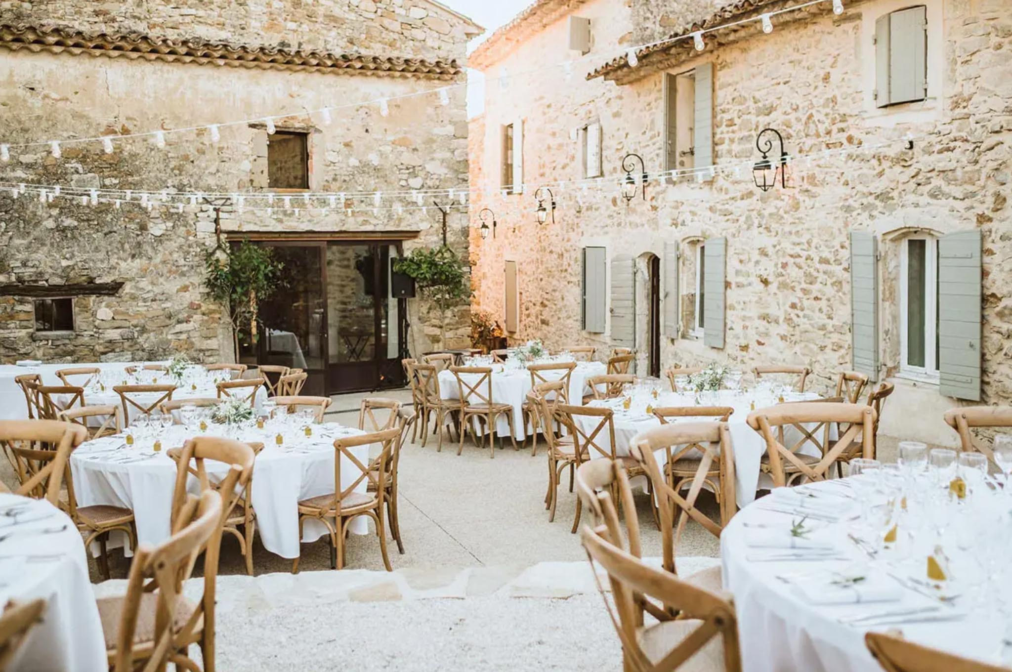 Round tables with greenery centerpieces under string lights in Provencal stone courtyard with sage shutters