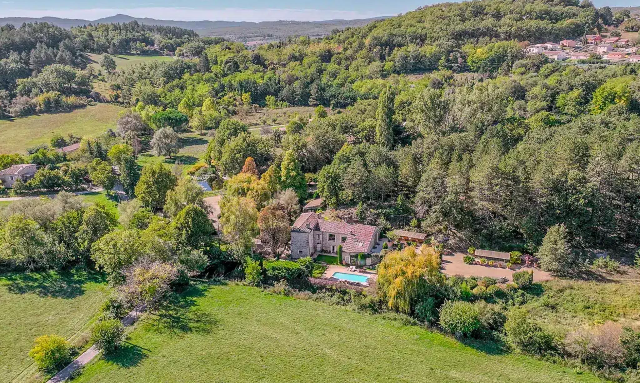 Aerial view of a stone country property with terracotta roof, swimming pool, and wooded estate in a rural valley