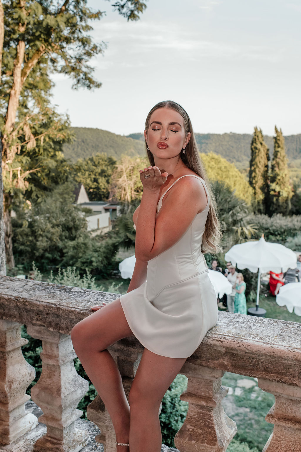 Bride in white mini dress blows kiss from stone terrace as guests mingle under umbrellas below at golden hour