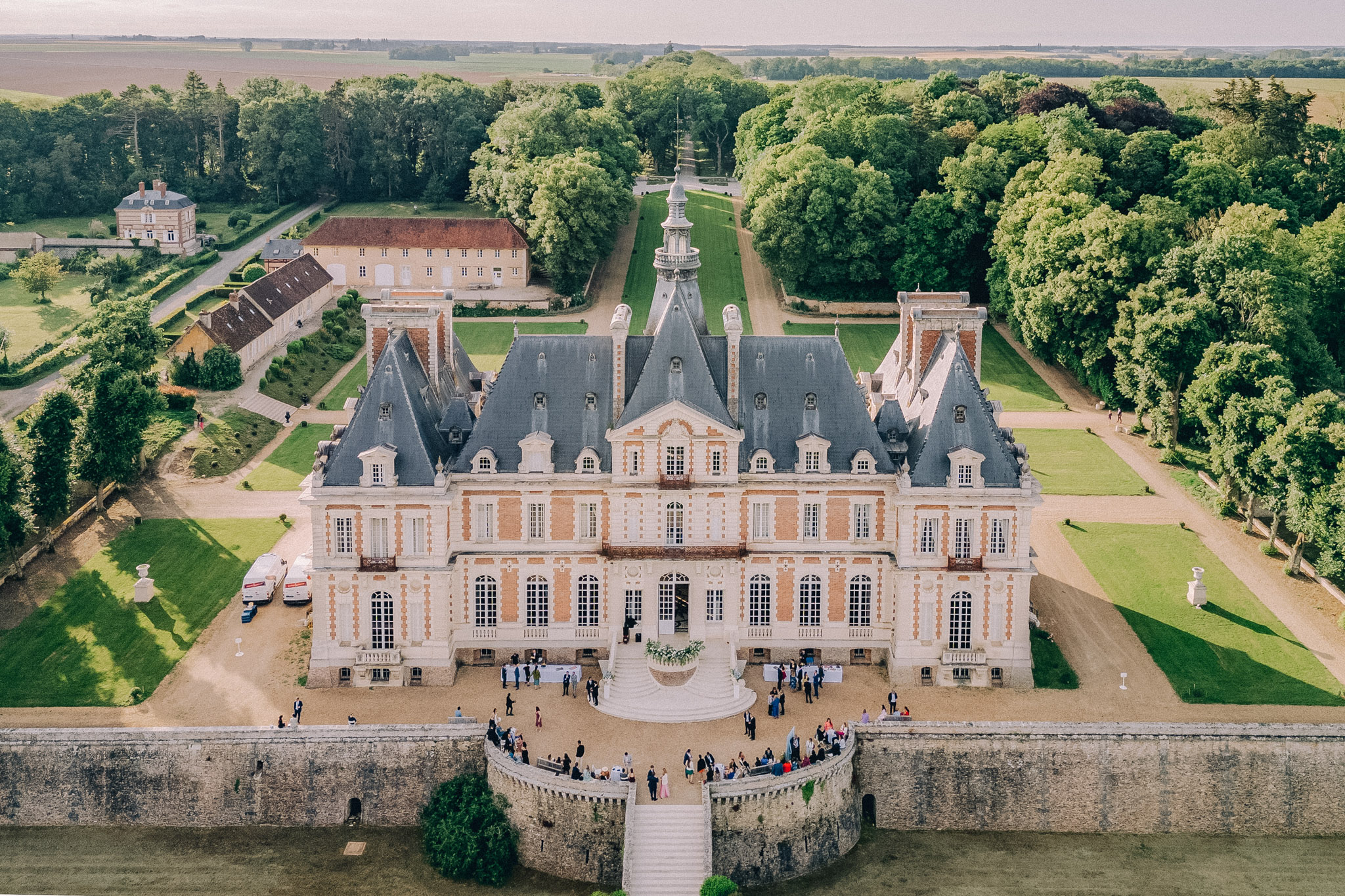 Aerial sixty guests mingling on forecourt of Louis XIII brick-and-stone chateau with mansard roof and formal tree-lined allee