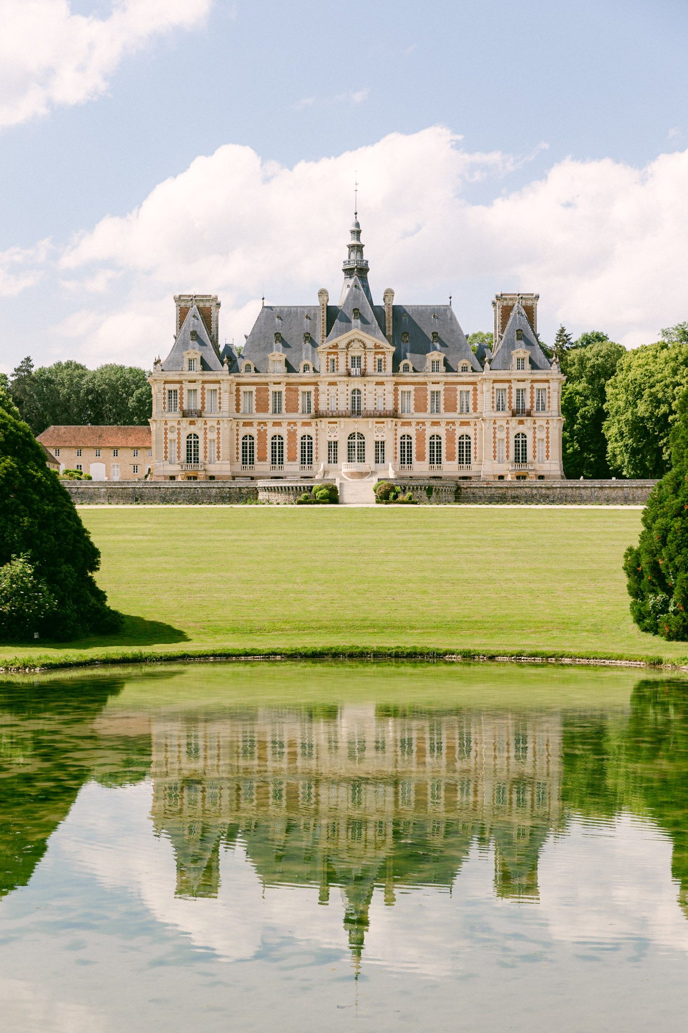17th-century chateau with mansard roof and spire reflected in formal rectangular pool across broad lawn