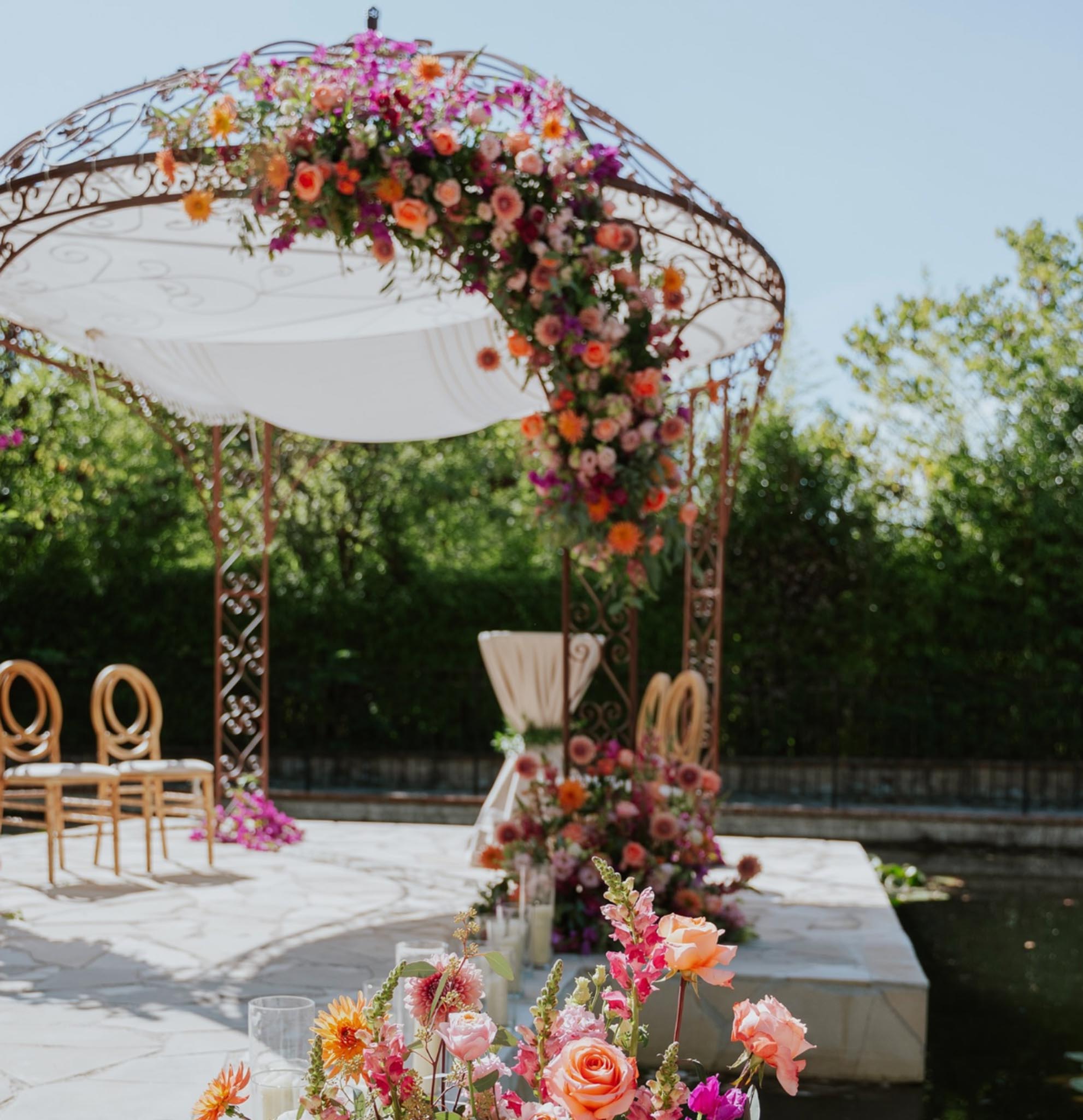 Wrought-iron gazebo draped with coral roses, magenta dahlias, and orange blooms on stone ceremony terrace