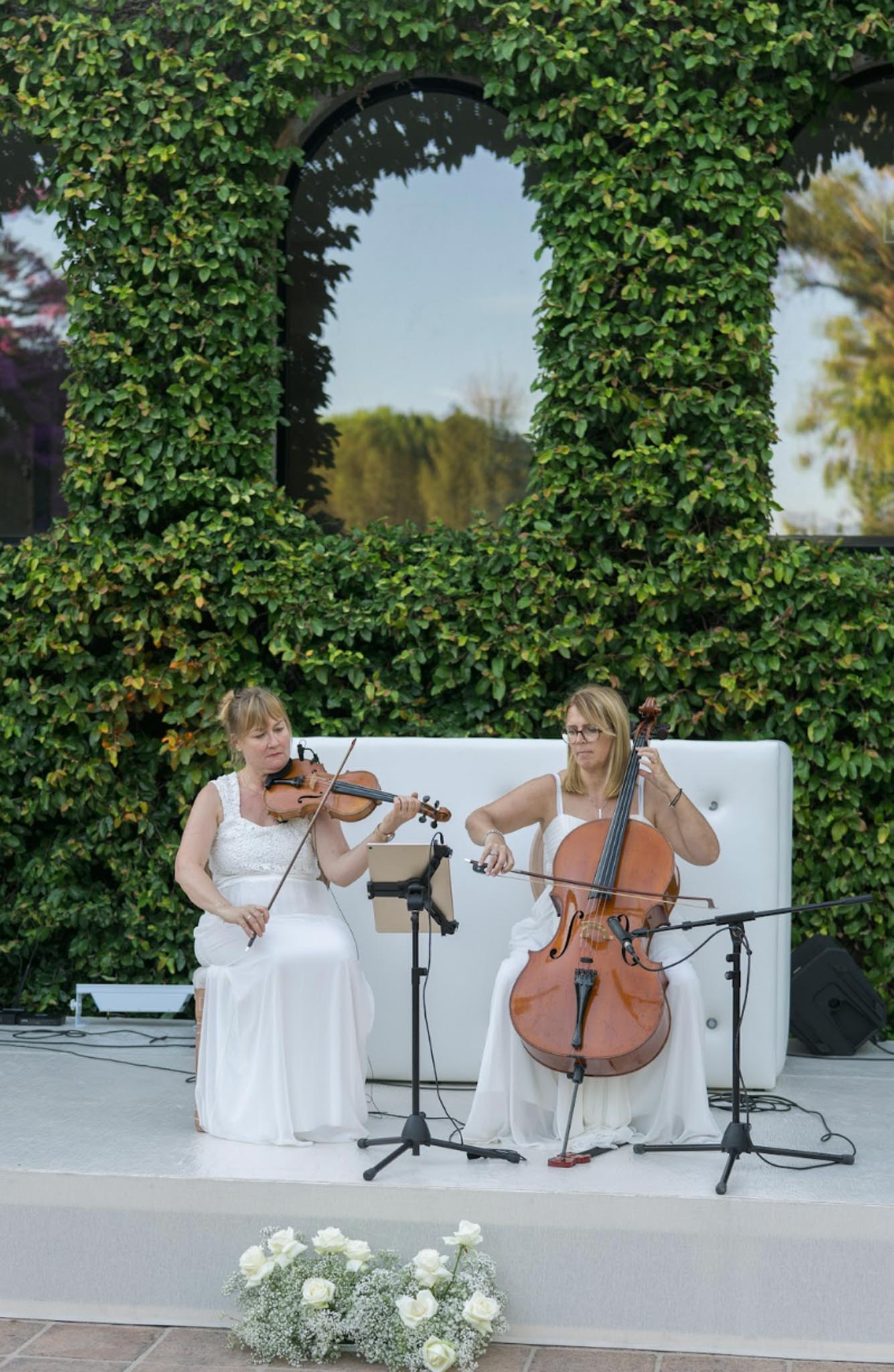 String duo in white gowns on tufted sofa performing before ivy wall with white rose arrangement at base