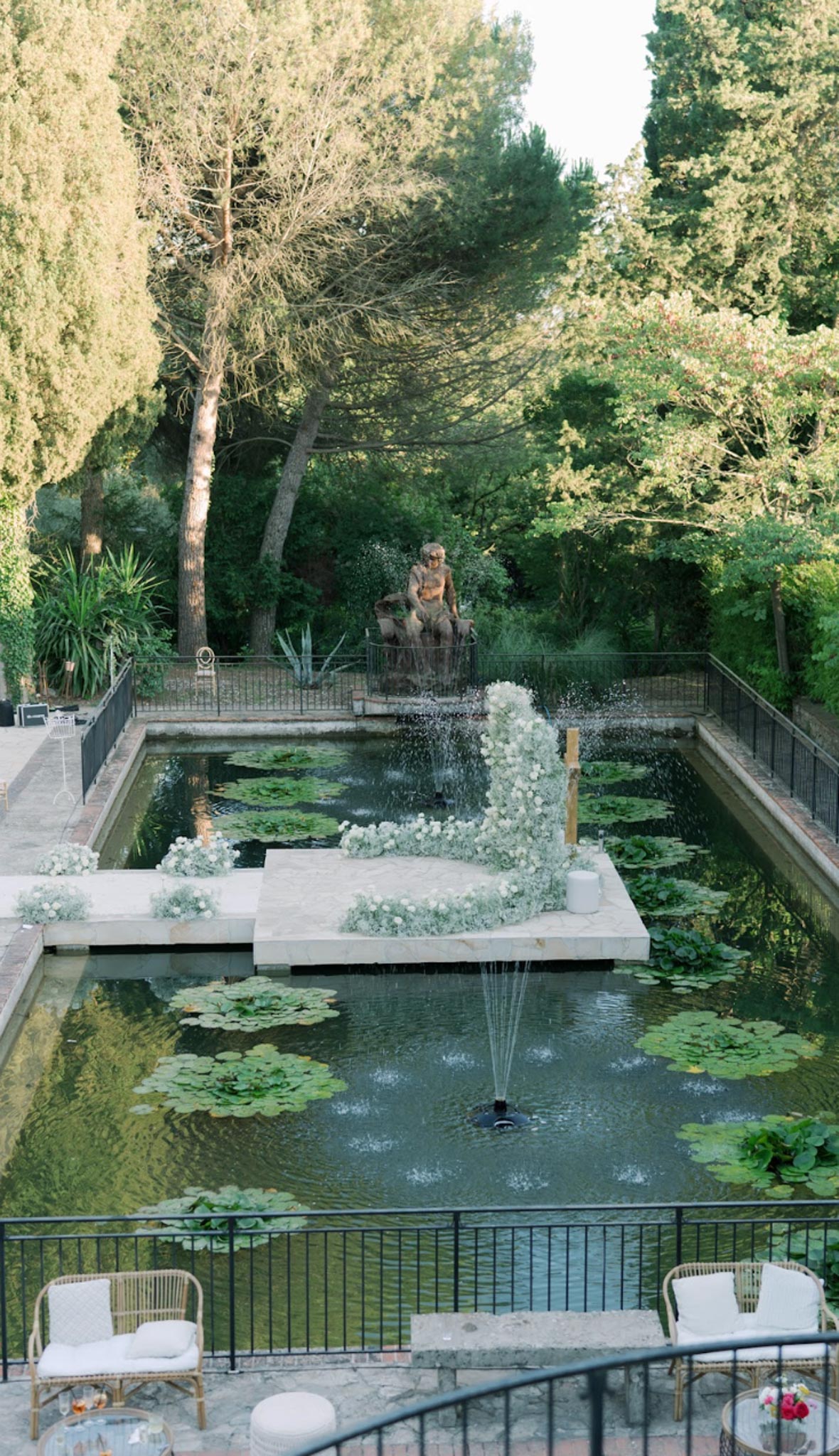 White crescent floral arch on floating platform in lily pond with rattan lounge chairs on terrace