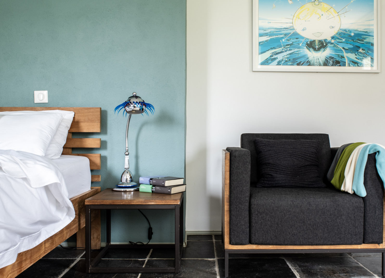 Modern bedroom with wooden bed, teal-green wall, charcoal armchair, chrome lamp, and dark slate floor