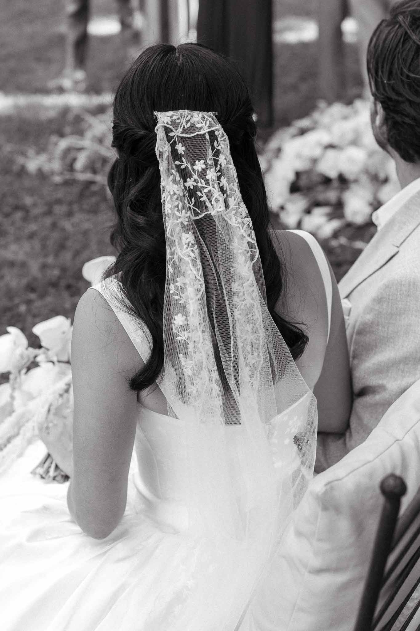 Black and white rear close-up of bride's floral-embroidered tulle veil beside groom during ceremony