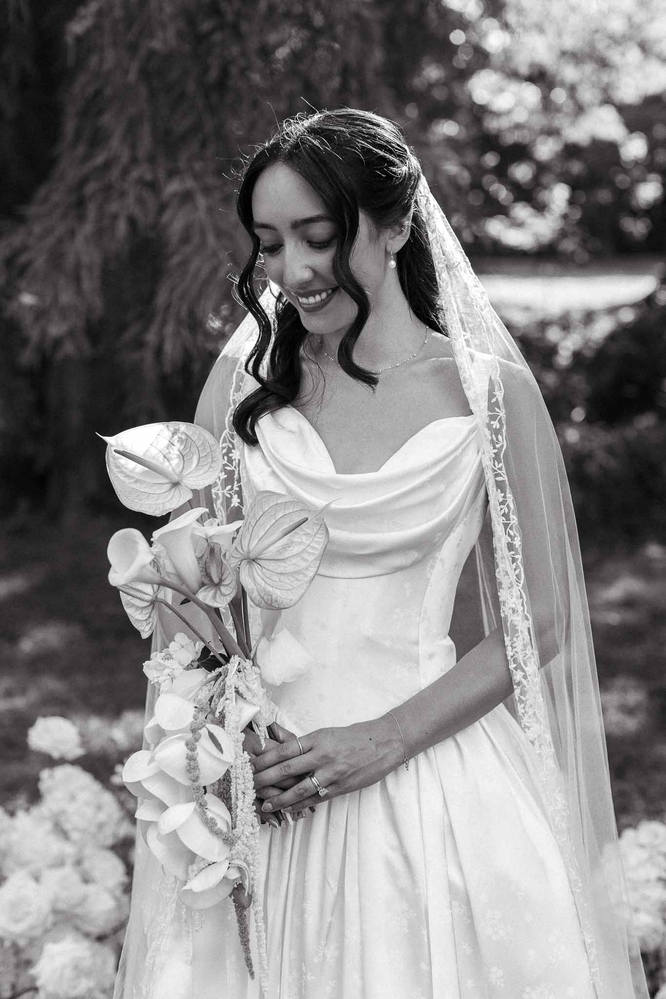 Black-and-white bridal portrait with off-shoulder satin gown, lace-trimmed veil, and trailing bouquet in garden