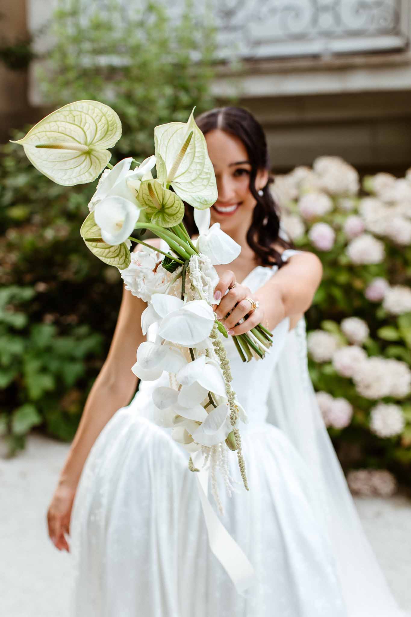 Bride holding bouquet of anthuriums, orchids, and calla lilies toward camera in stone courtyard garden