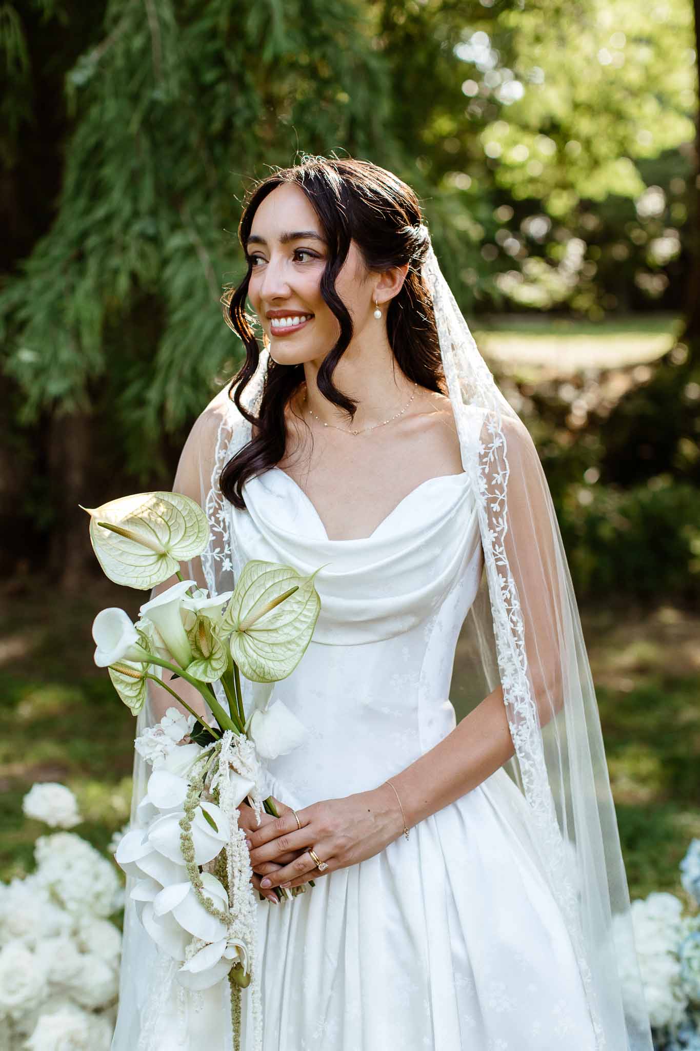 Bride in ivory satin gown holding cascading bouquet of white anthuriums calla lilies and orchids in garden setting