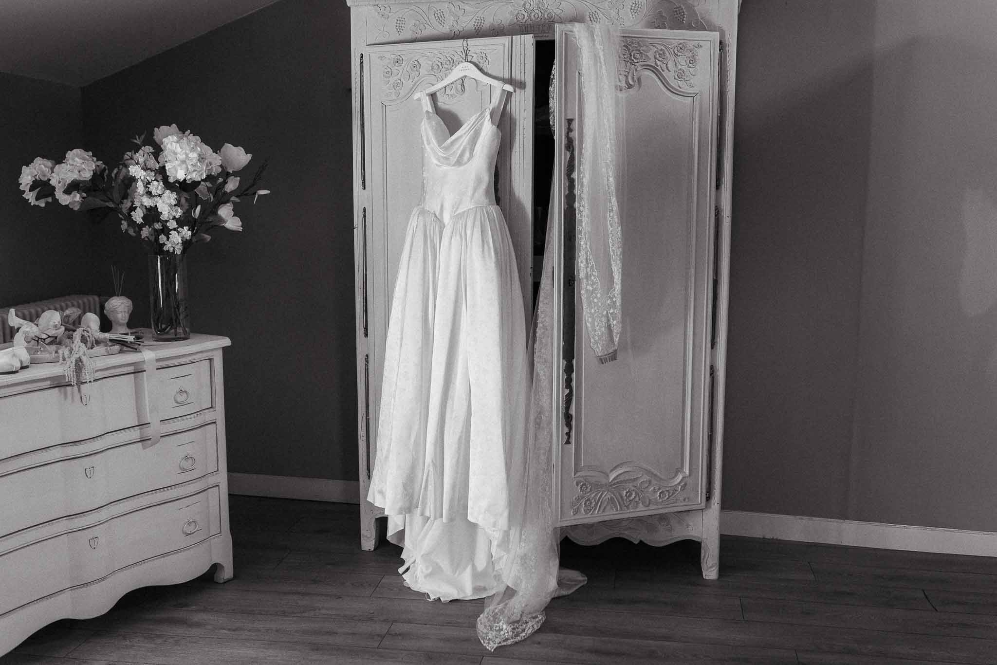 Black and white of cowl-neck satin gown and lace veil hung on carved armoire beside hydrangea arrangement