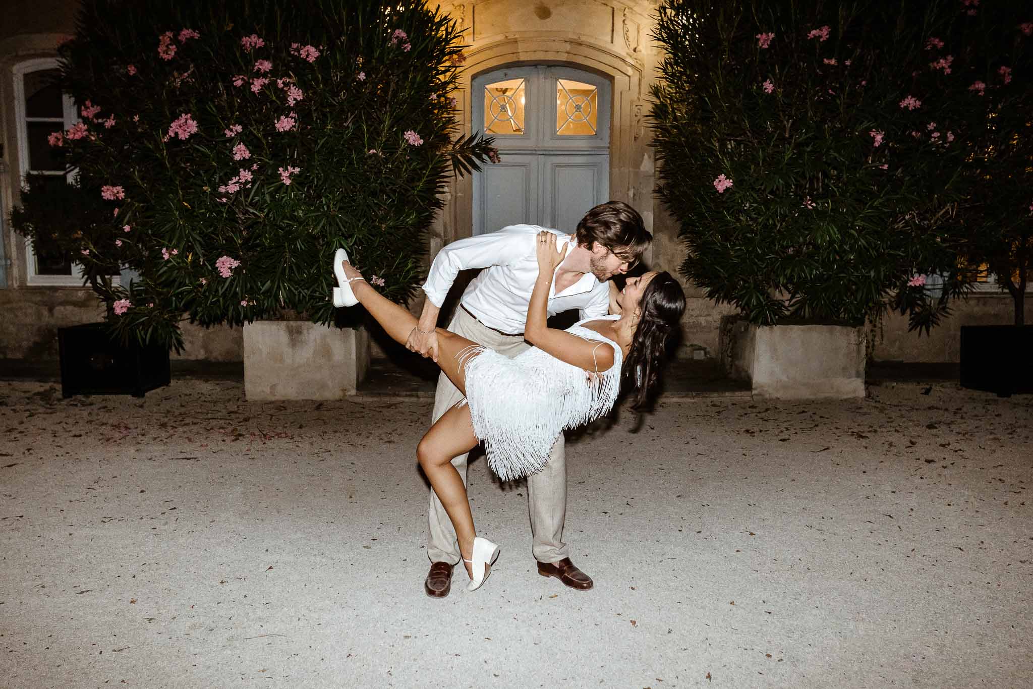 Groom dipping bride in white fringe mini dress for kiss during evening courtyard reception