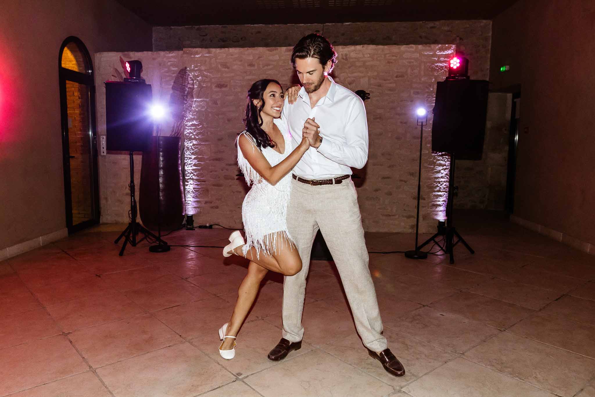 Bride in white fringe mini dress mid-dance move with groom in linen trousers inside stone-walled barn with pink uplighting