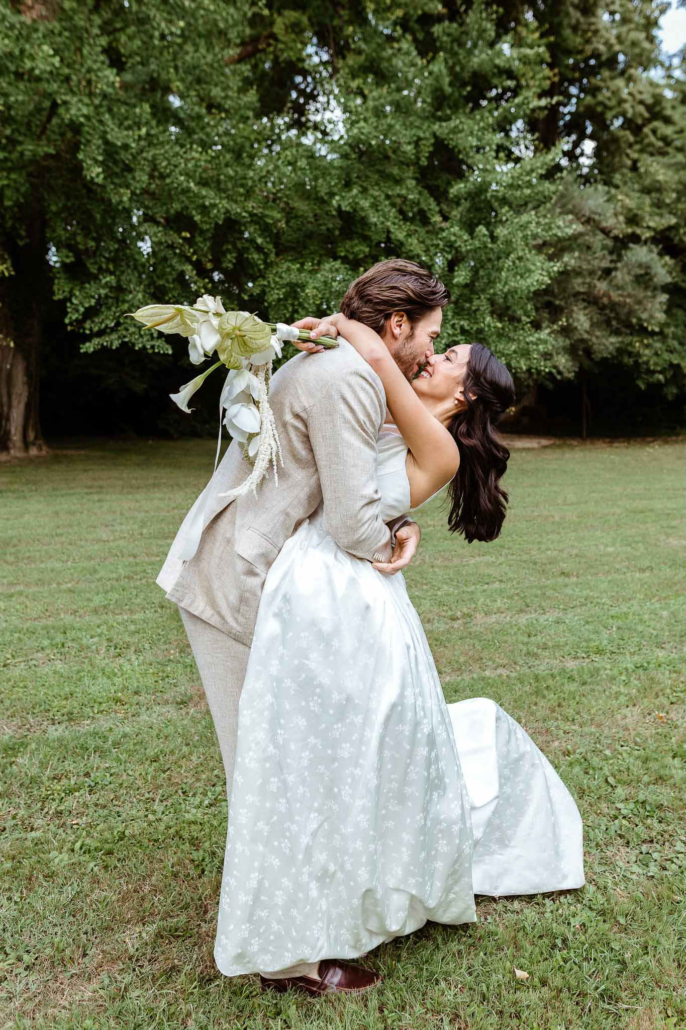 Groom dipping bride for a kiss on garden lawn, bride holding white anthurium and calla lily bouquet with trailing amaranthus
