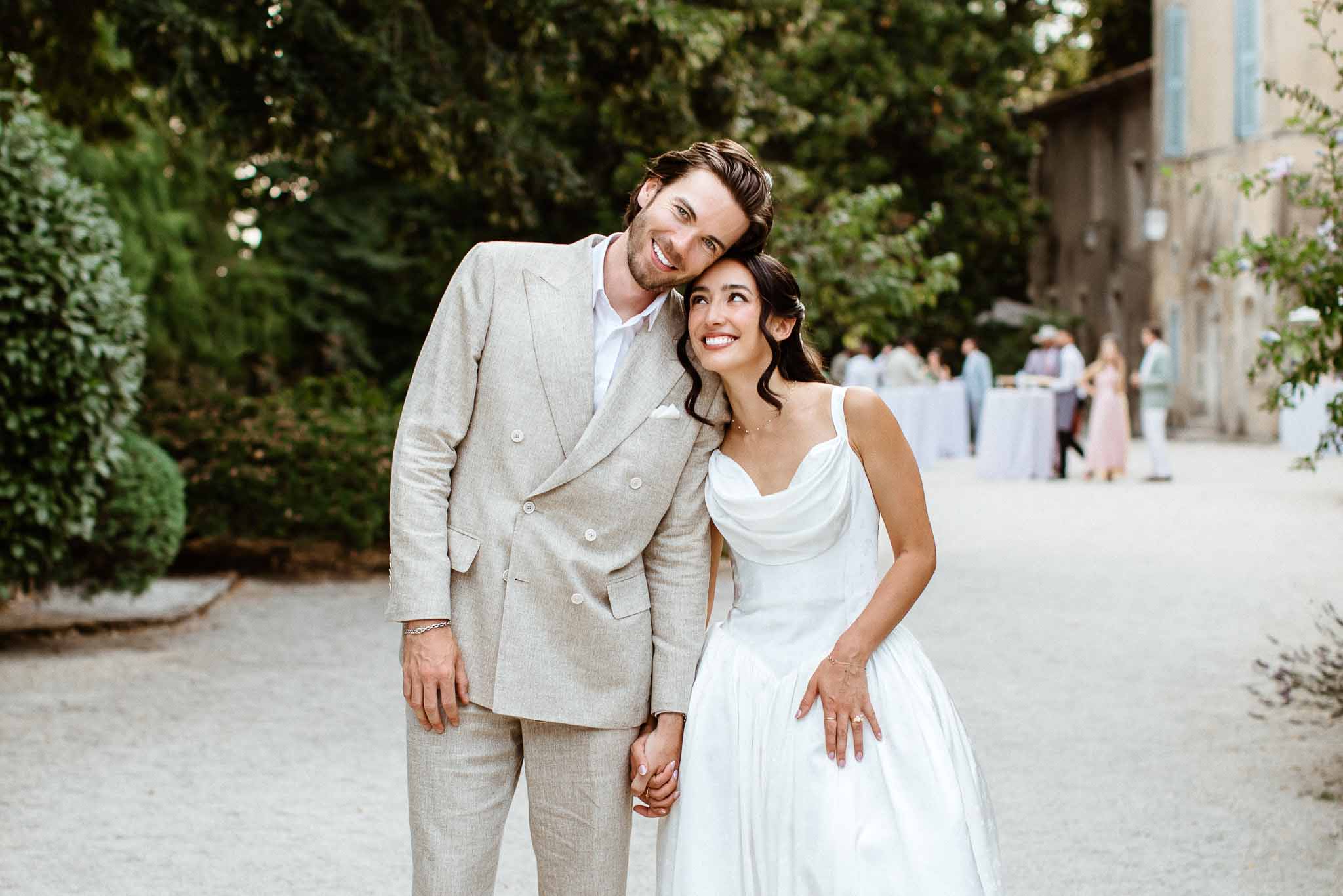 Bride in white satin cowl-neck gown and groom in beige linen suit holding hands on gravel courtyard