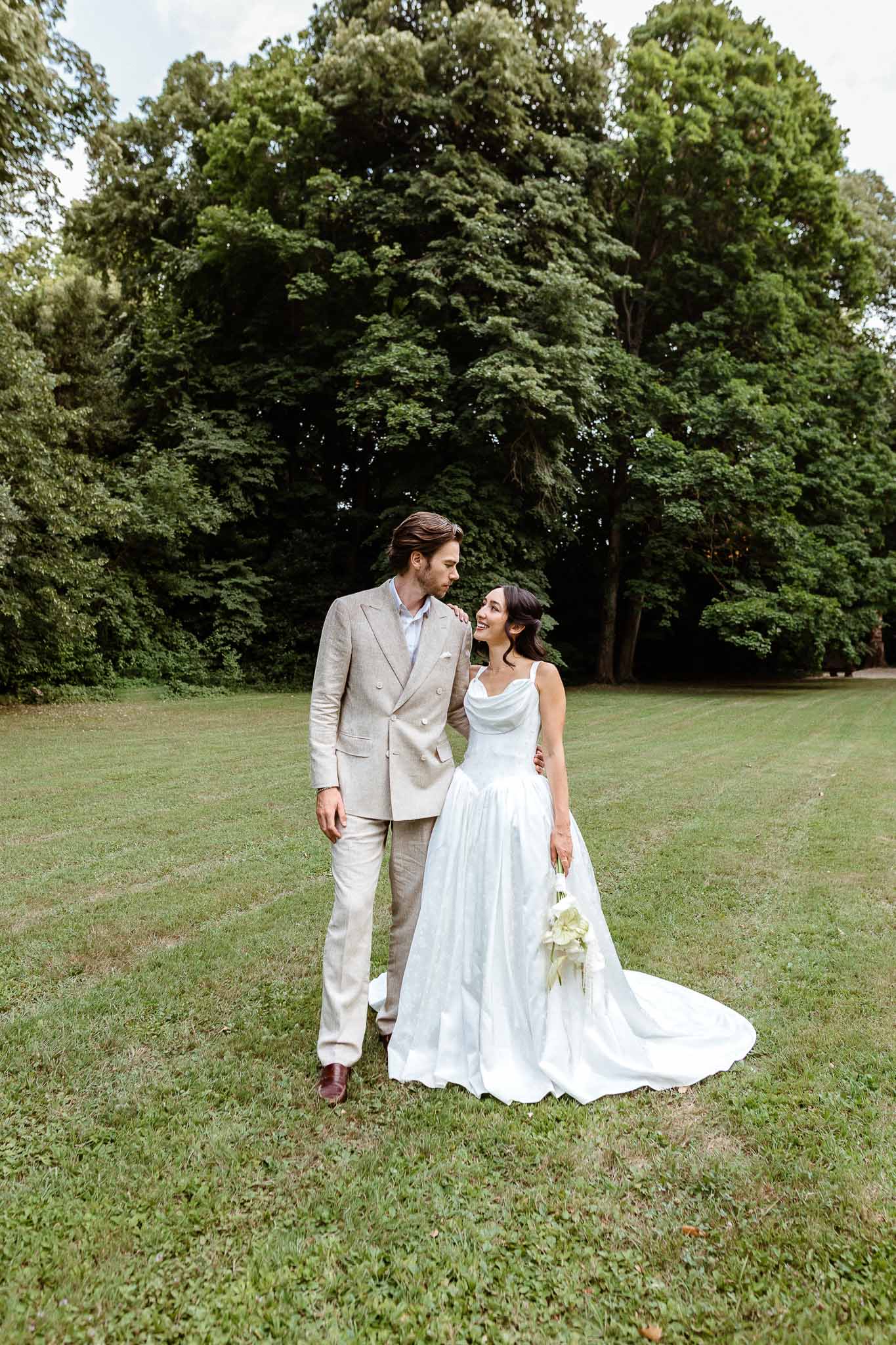 Bride in white satin cowl gown with calla lily bouquet and groom in oatmeal linen suit on lawn