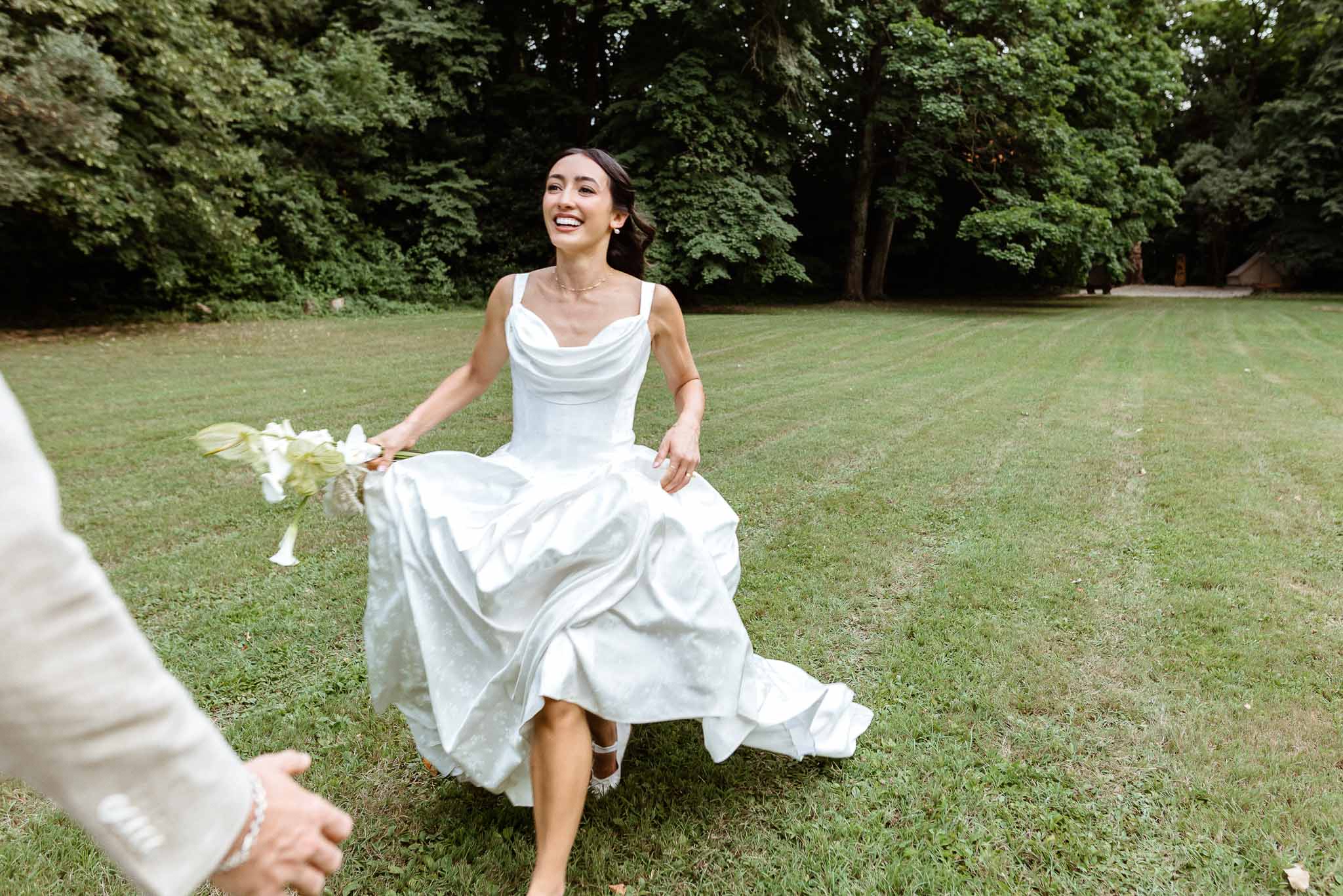 Bride in white satin cowl-neck gown laughing and running across lawn holding calla lily bouquet toward groom