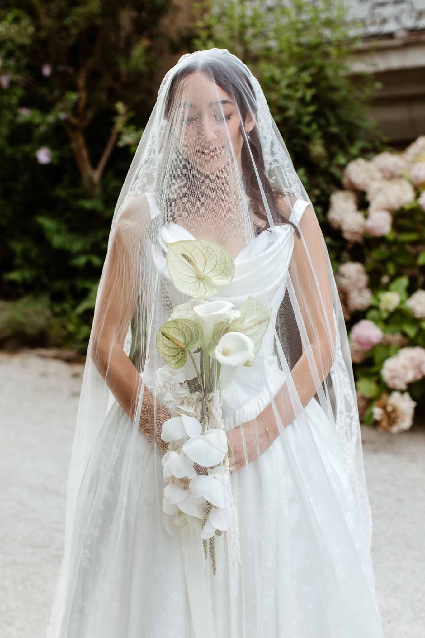 Bride in ivory satin cowl-neck gown and lace-edge cathedral veil holding white anthurium and calla lily bouquet