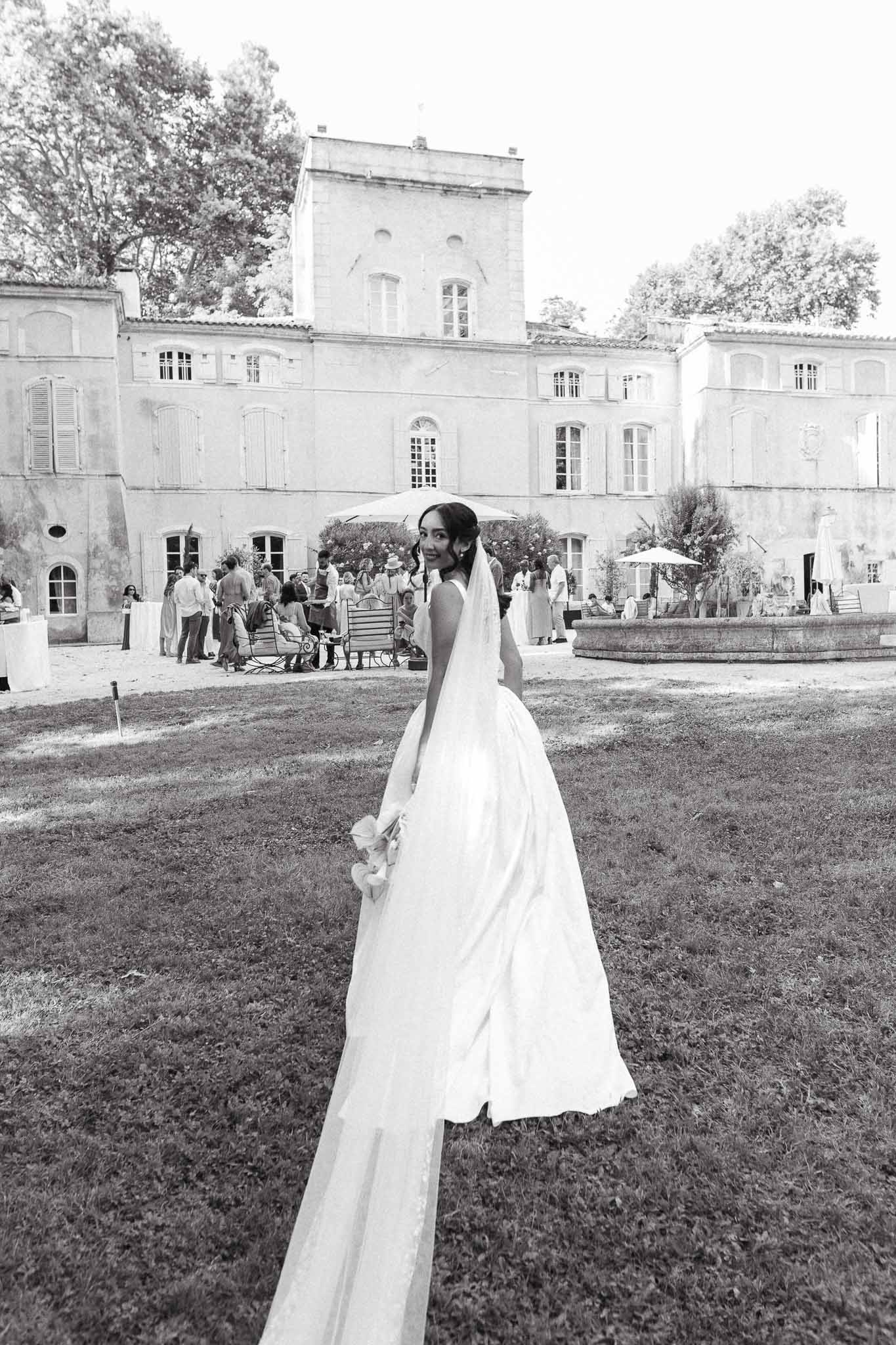 Black and white bride looking over shoulder with ball gown train and veil as guests mingle by chateau fountain