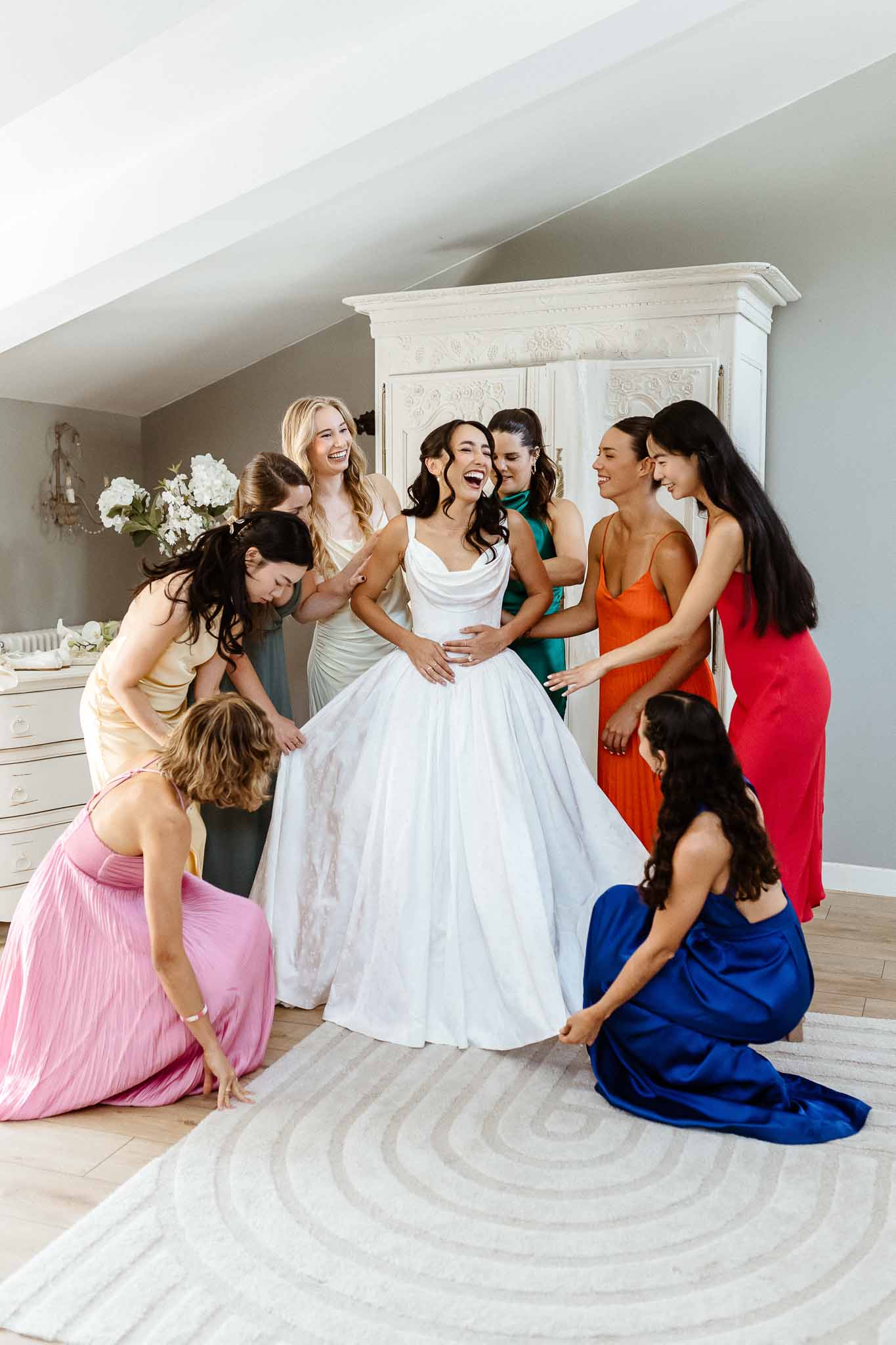 Bride in white ballgown laughing with eight bridesmaids in mismatched colourful satin dresses