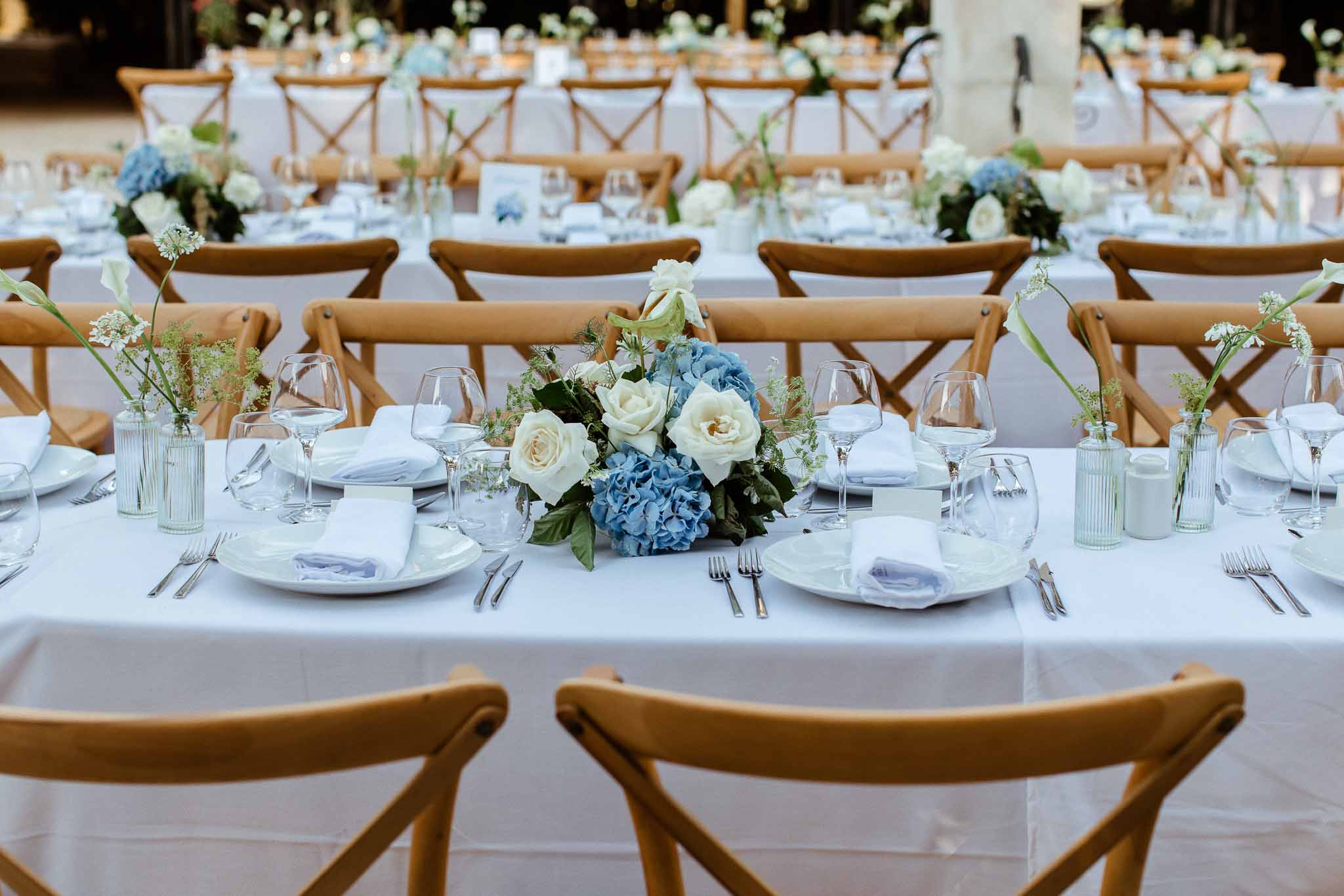 Reception table with dusty blue hydrangea centerpiece, ivory roses, blue napkins, and cross-back chairs