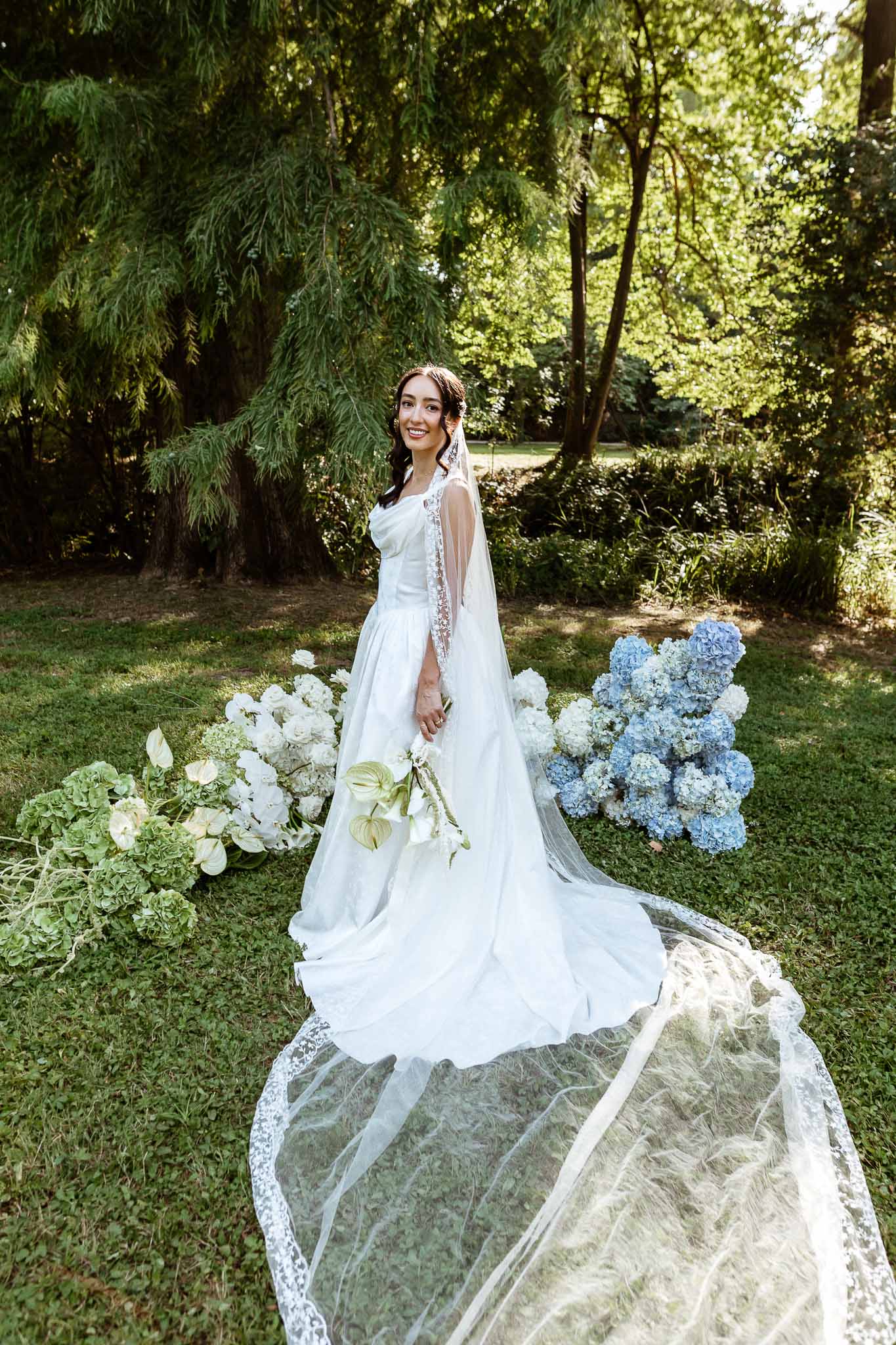 Bride in white A-line gown with cathedral lace veil on garden lawn beside white and blue hydrangea arrangements