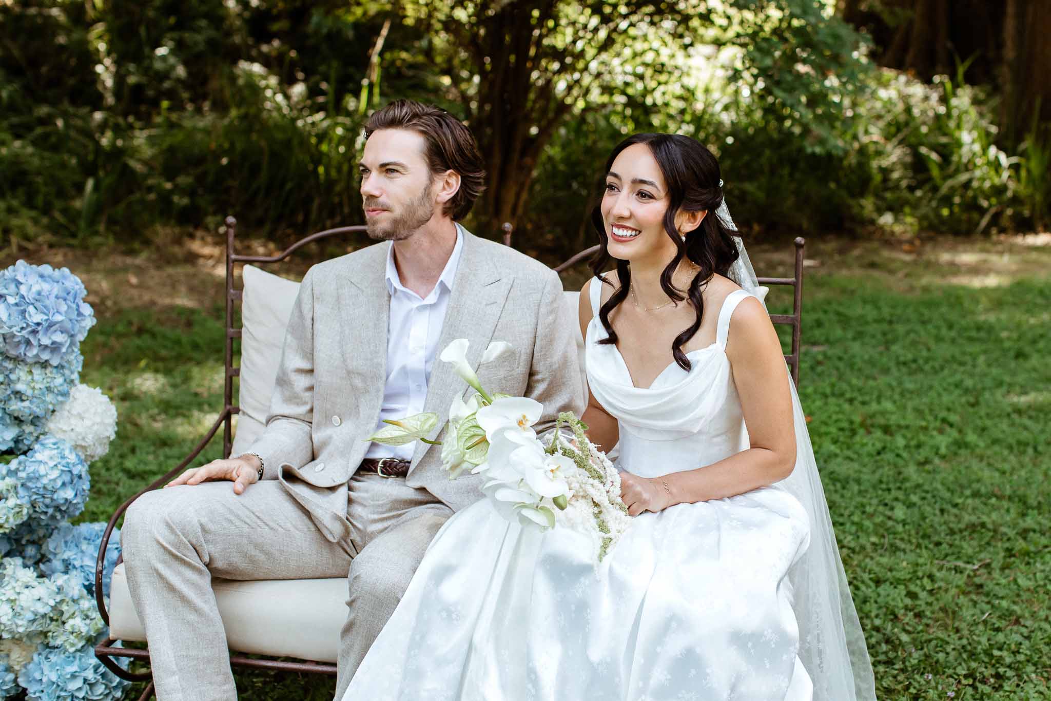 Couple seated on iron bench with calla lily bouquet beside blue and white hydrangea ceremony arrangement