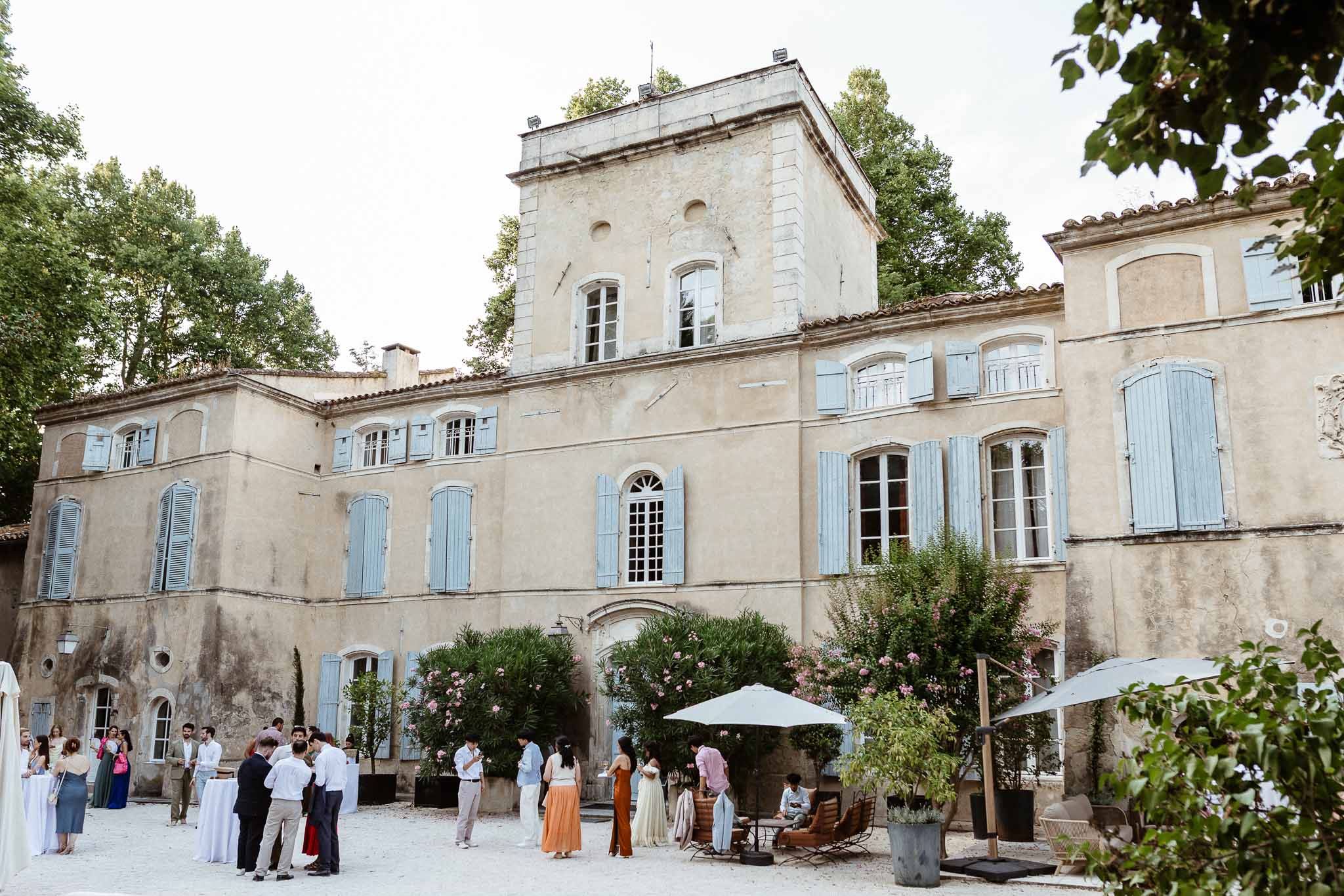 Guests mingling in gravel courtyard of Provencal bastide during cocktail hour with rattan chairs and umbrellas