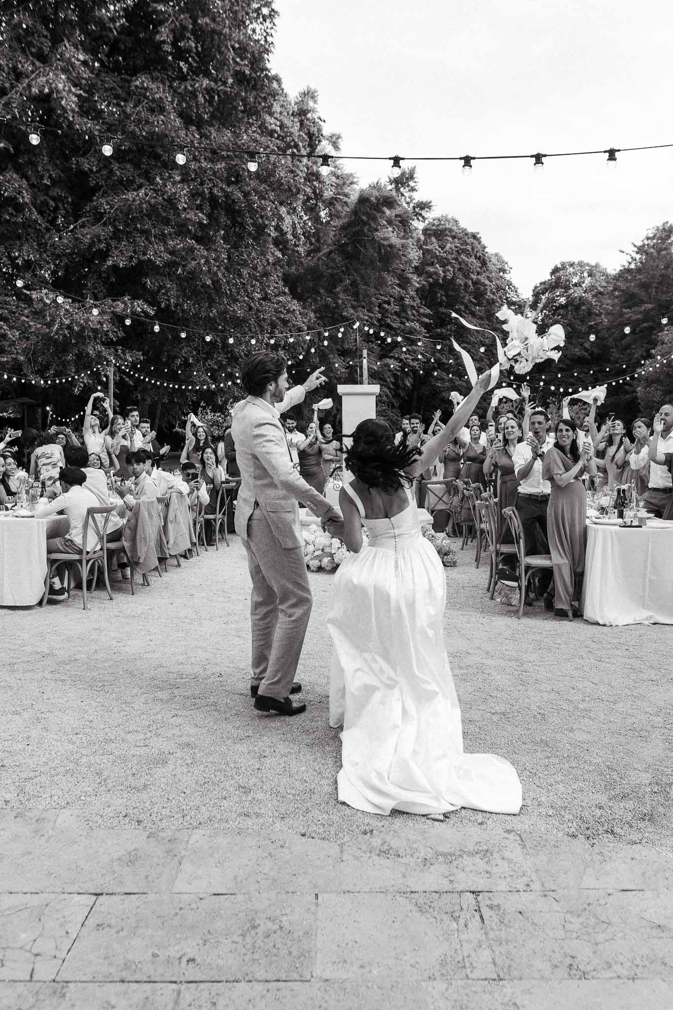 Black-and-white shot of couple entering outdoor reception as guests cheer under festoon lights on terrace