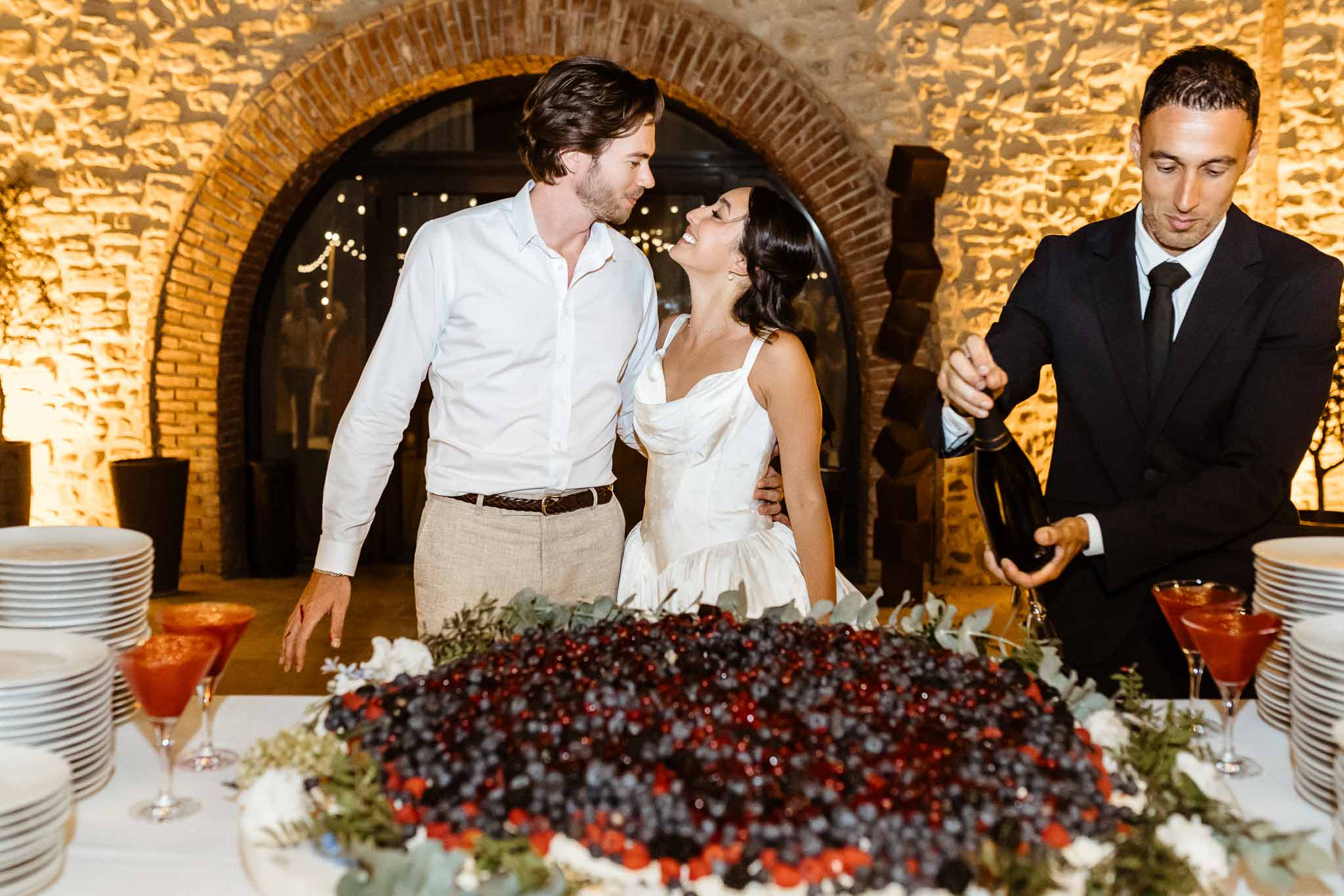 Couple smiles at each other during cake cutting beside a berry-covered wedding cake inside a stone venue with fairy lights