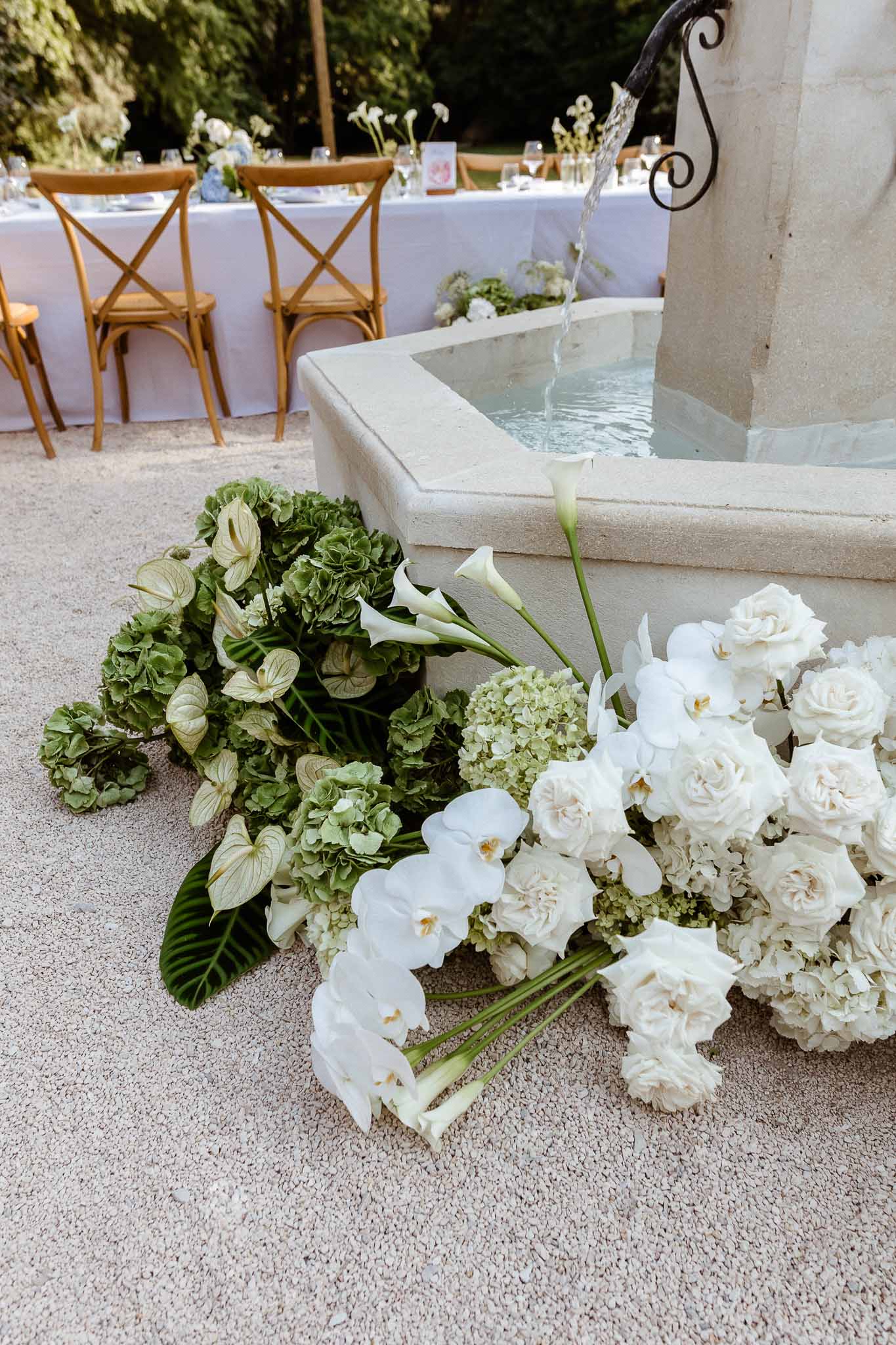 Stone fountain with white orchid and rose floral arrangement at base, reception table with cross-back chairs in background